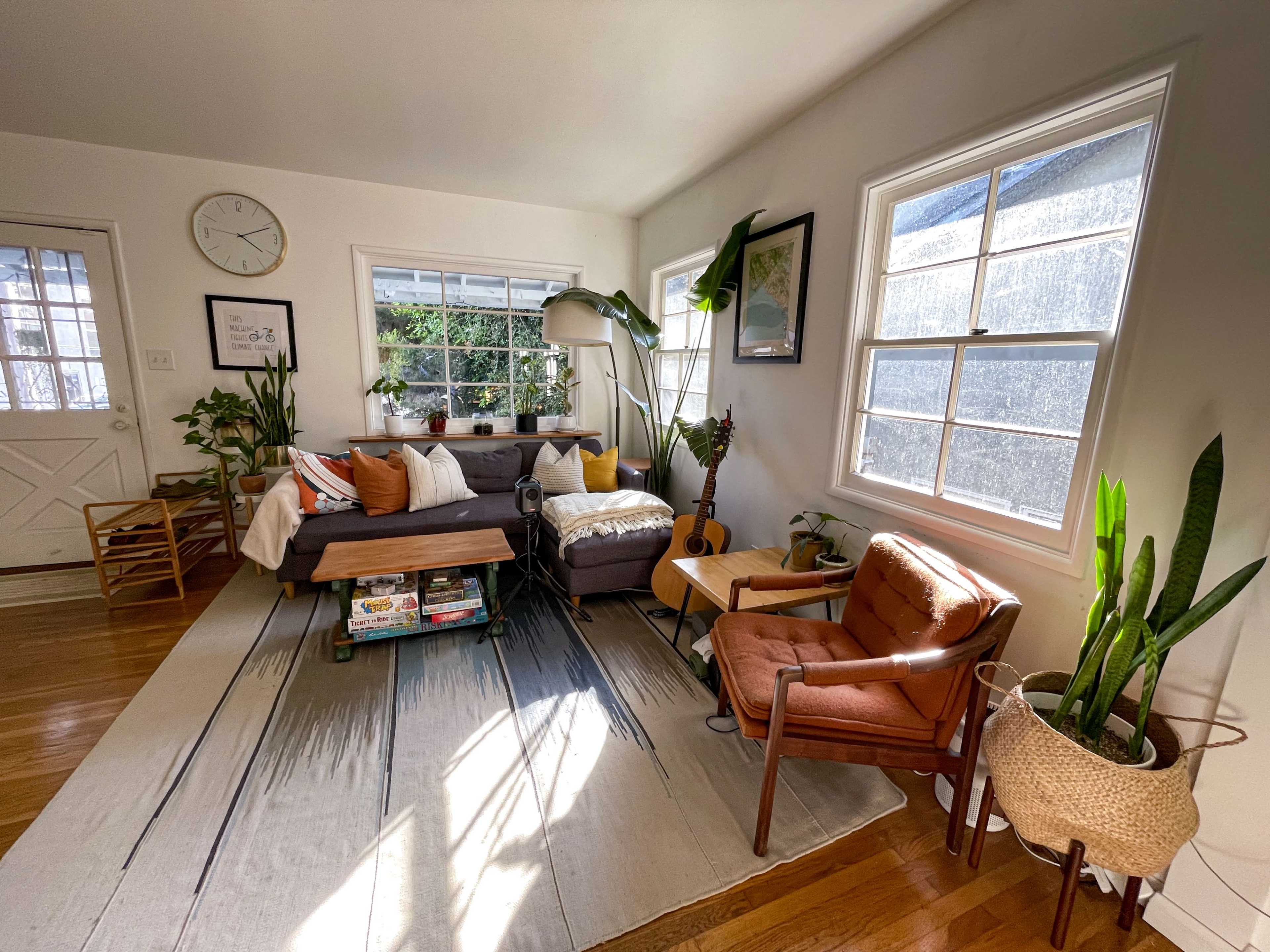 A bright living room features a sofa, a wooden coffee table, an orange armchair, and several plants by the windows.