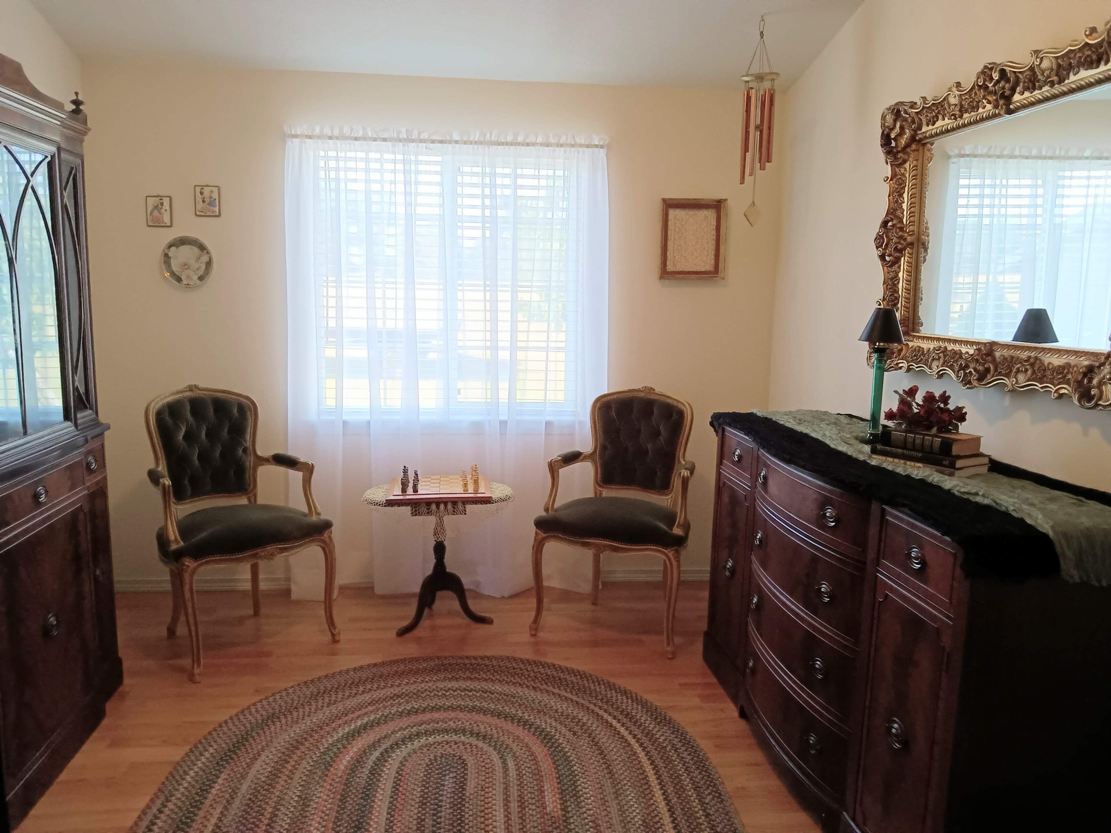 The image shows a cozy room with two upholstered chairs, a small table between them, a decorative mirror on the wall, and a dresser adorned with a plant.