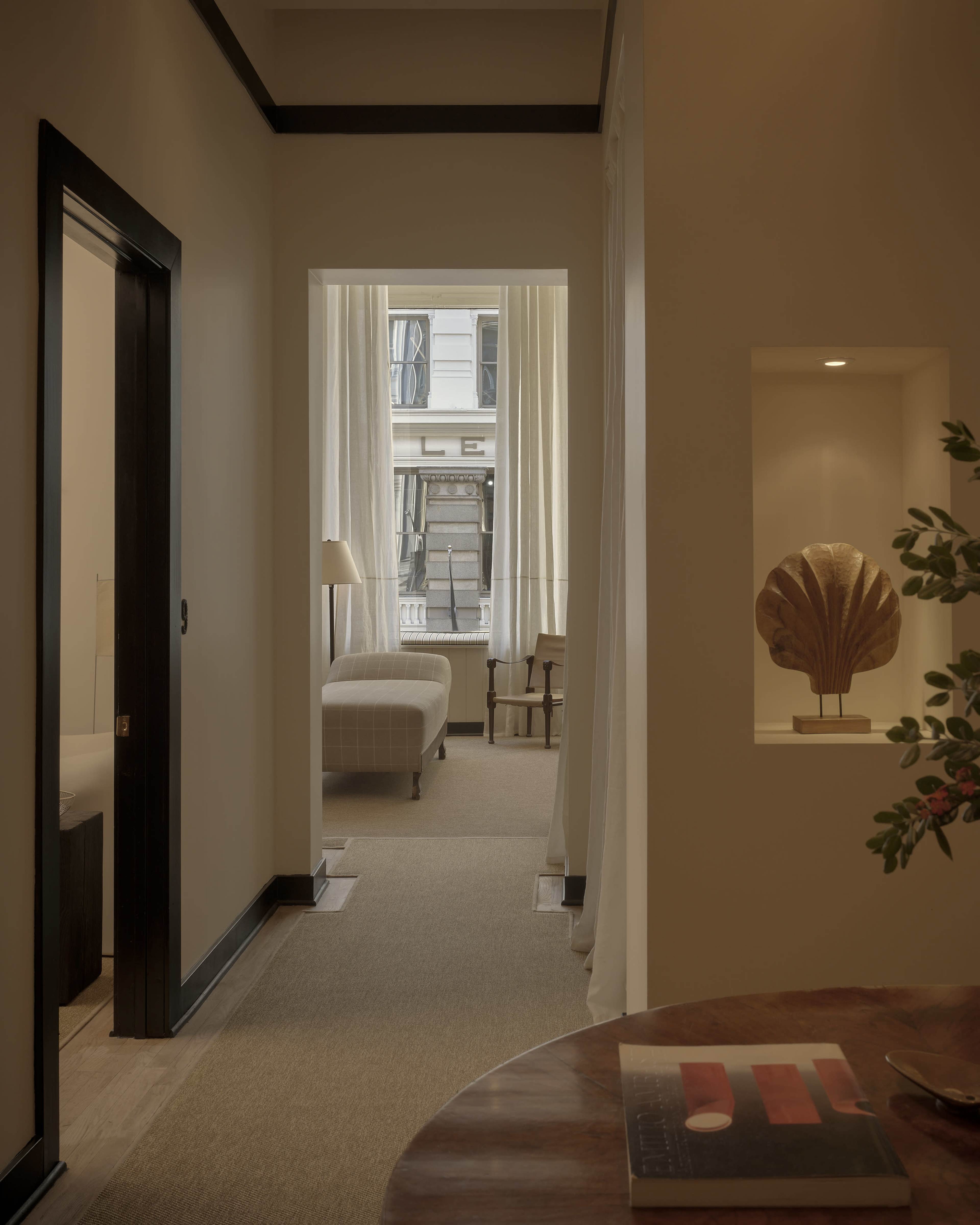 The image shows a softly lit corridor leading to a room with a window, a chair, and a decorative element on the wall.