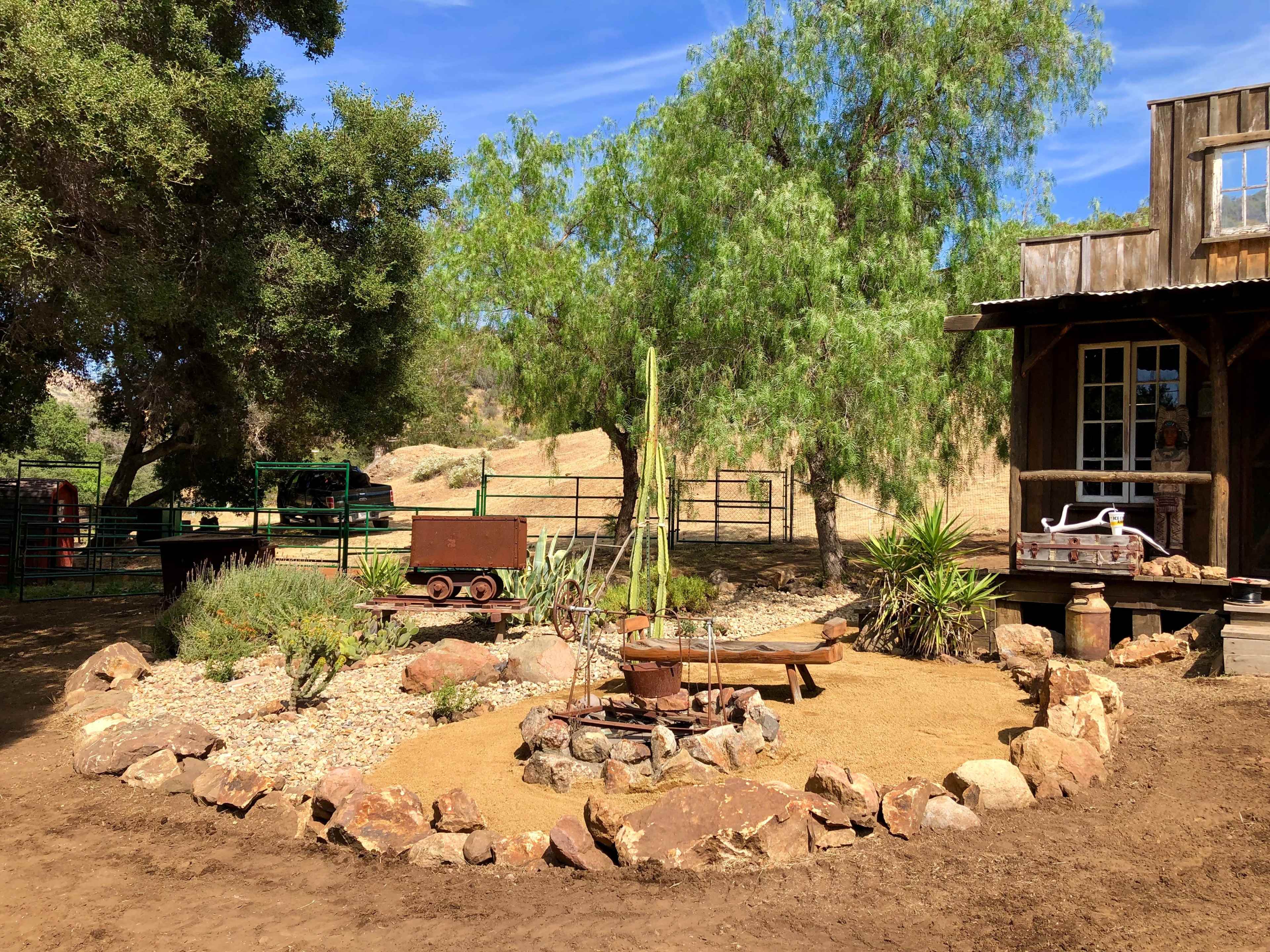 A small outdoor area with a fire pit surrounded by rocks and plants, located next to a rustic wooden structure and trees.