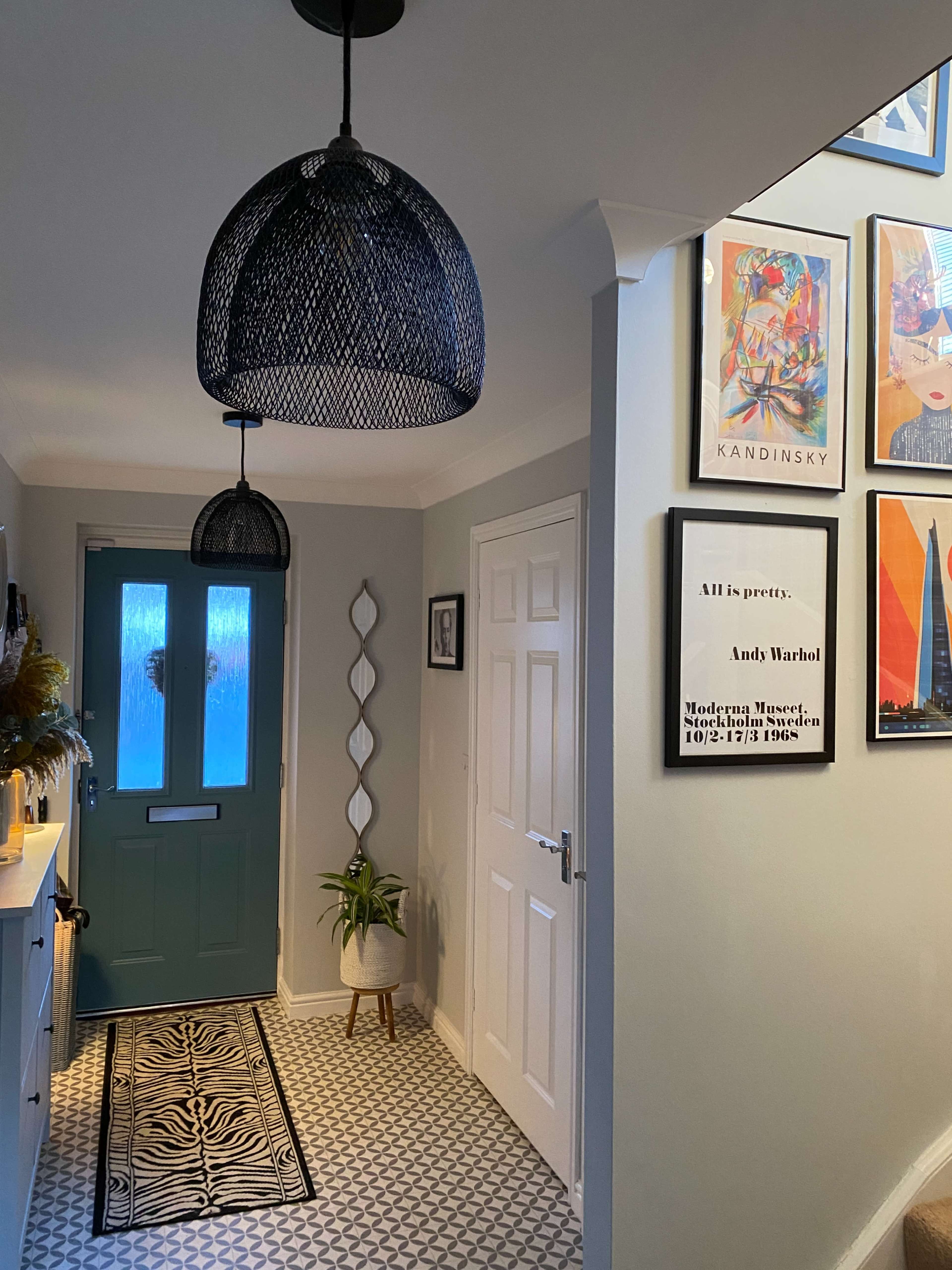 A hallway with two decorative pendant lights, a green door, framed artwork on the walls, and a runner rug on a patterned floor.