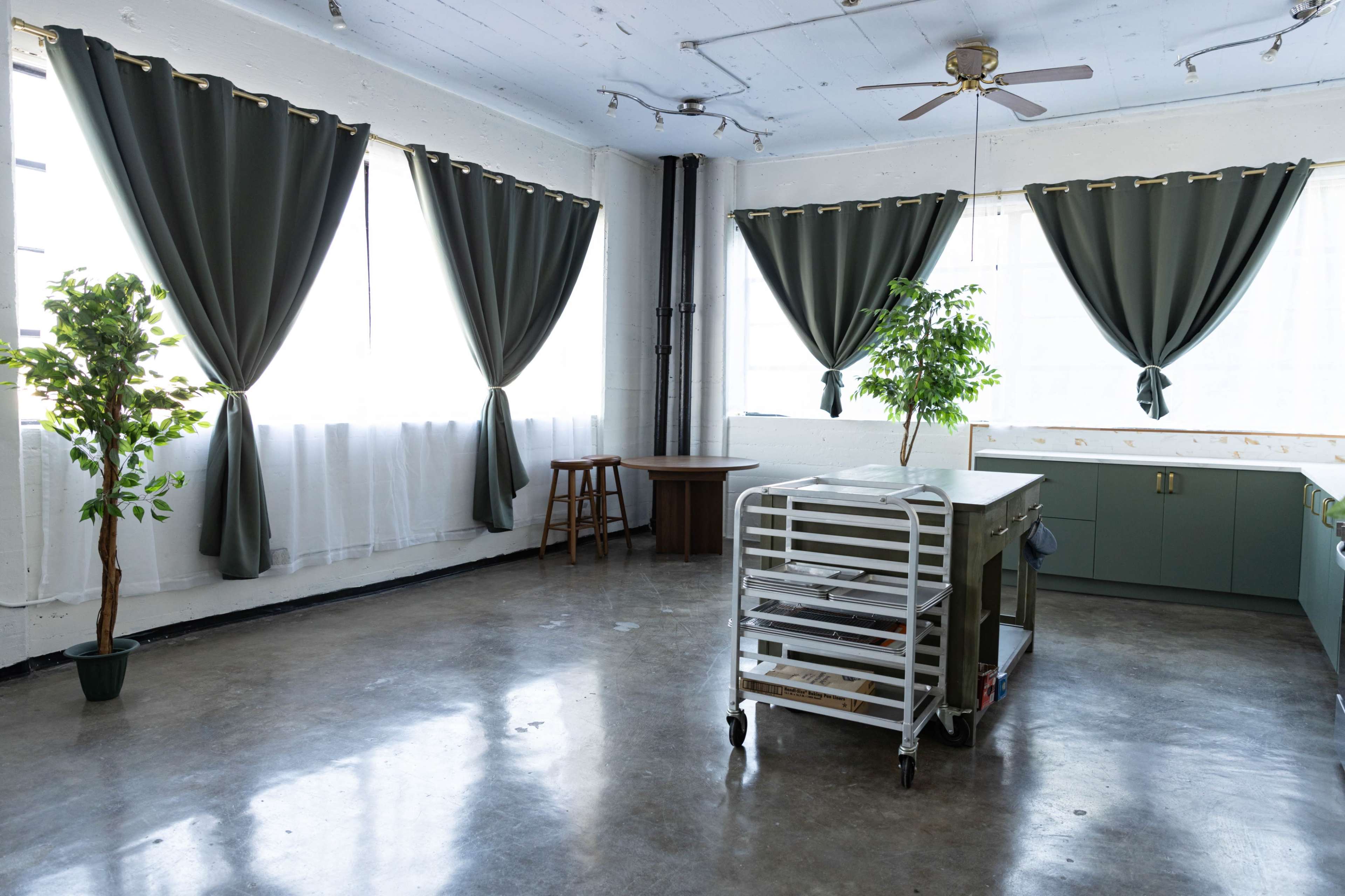 The image shows a spacious room with large windows covered by long curtains, a plant, a rolling cart, and a table.