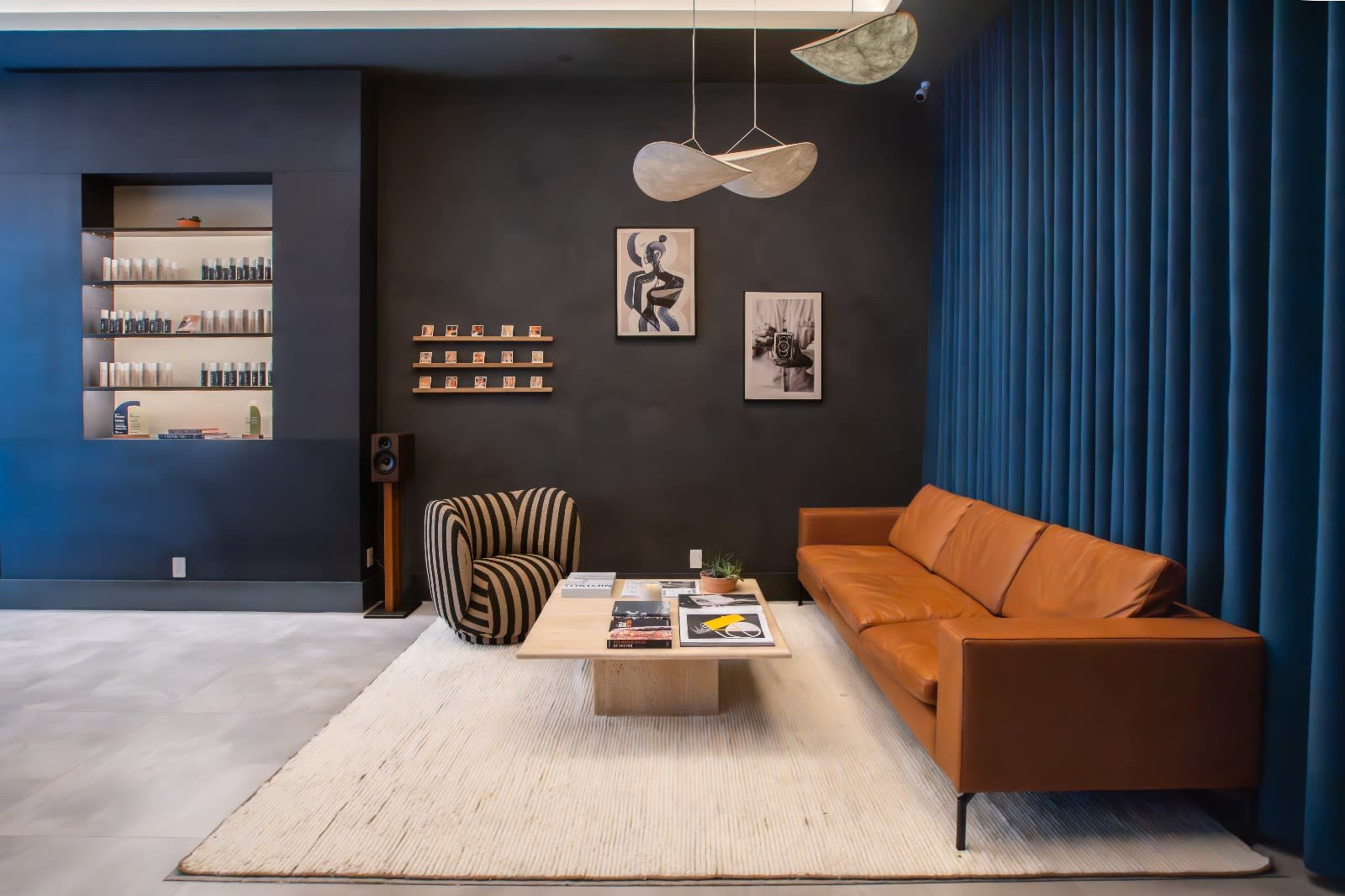 The scene features a modern lounge area with a brown leather sofa, a striped chair, a wooden coffee table, and a dark wall displaying artwork and shelves.