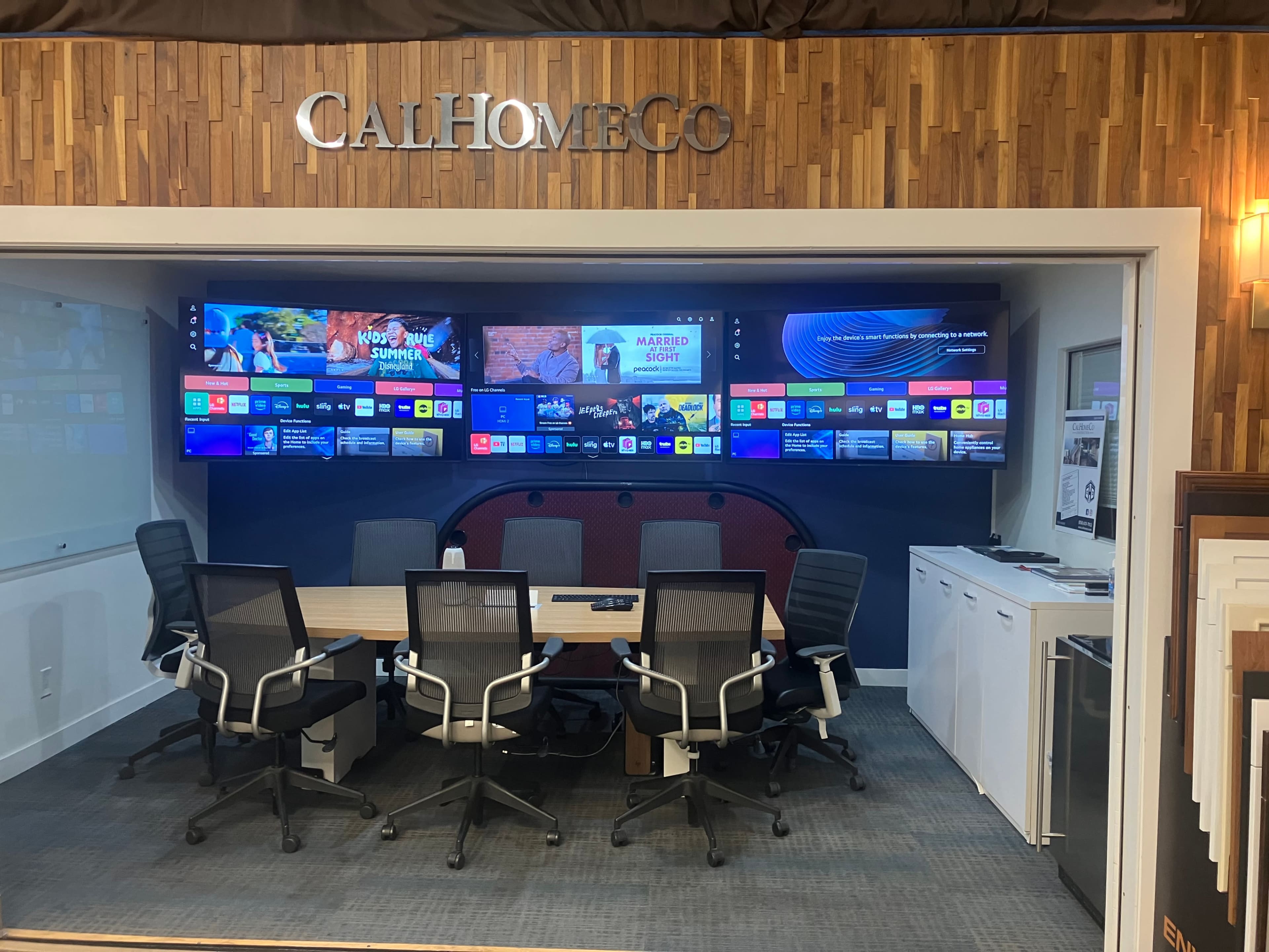 The image shows a modern conference room with a large table surrounded by ergonomic chairs, multiple screens displaying information, and wooden paneling on the walls.