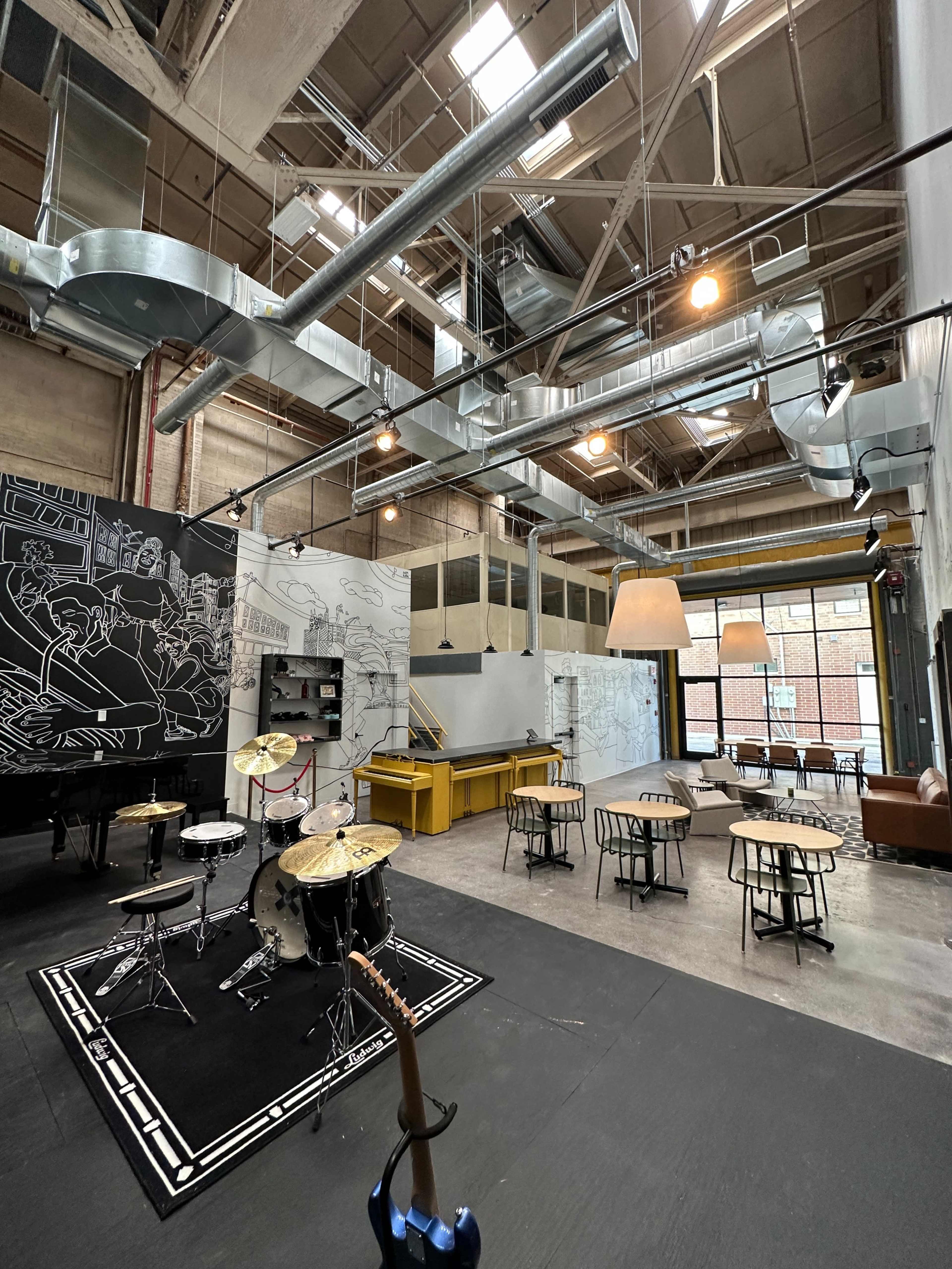 An industrial-style venue with a stage for music, a seating area with tables, and large windows letting in natural light.