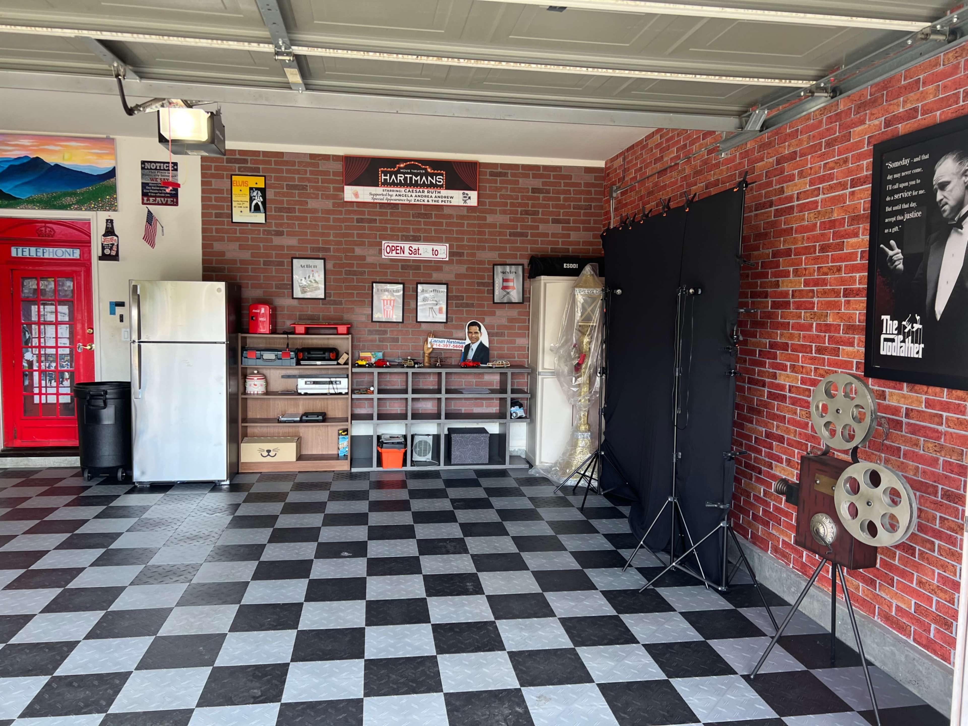 A garage with a checkered floor, a brick wall backdrop, and various items including a refrigerator, a shelf with storage, and a film projector.