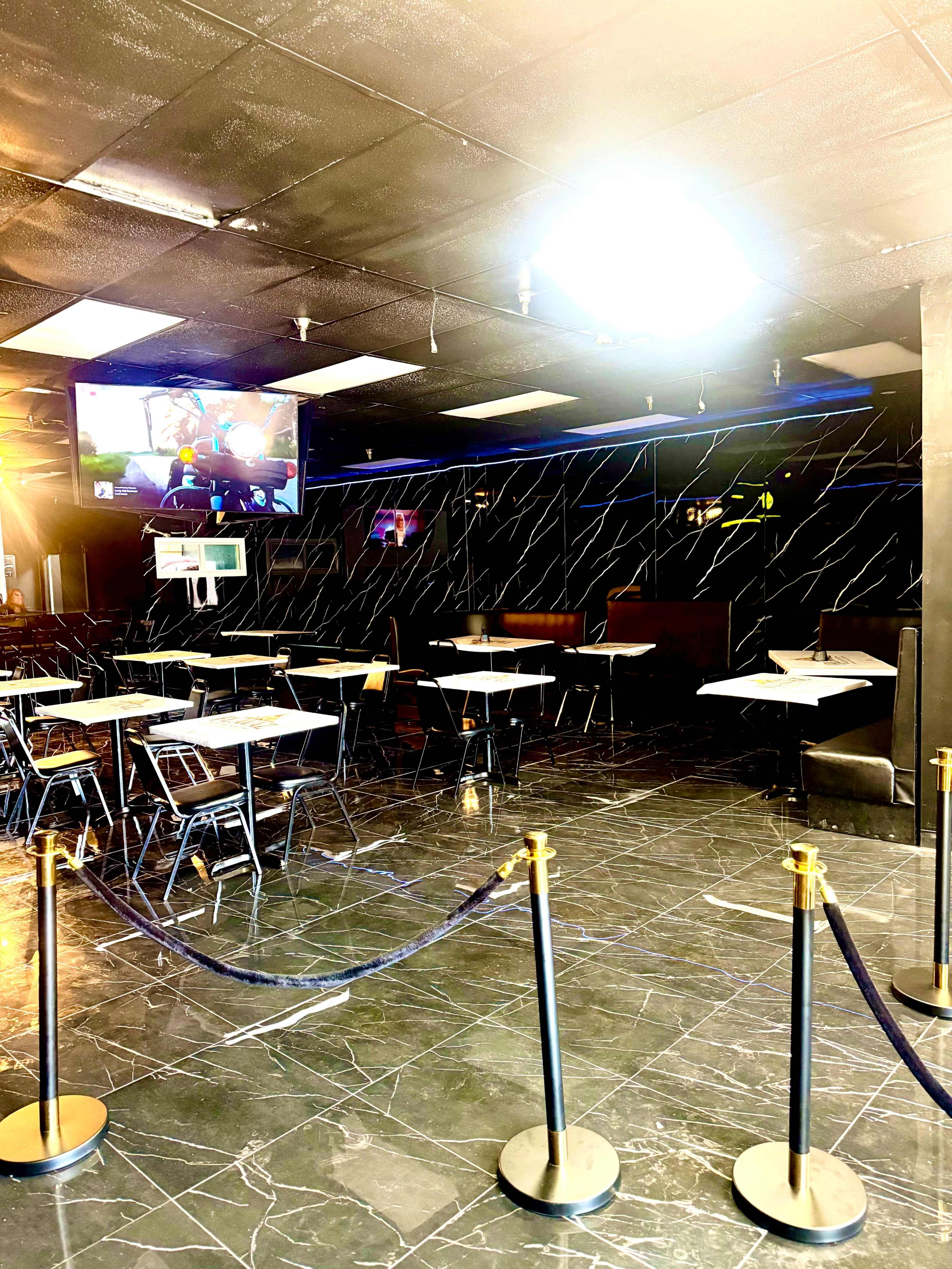 The image shows an empty indoor dining area with tables and chairs arranged around a black and white marble floor, and a television mounted on a wall displaying a game.