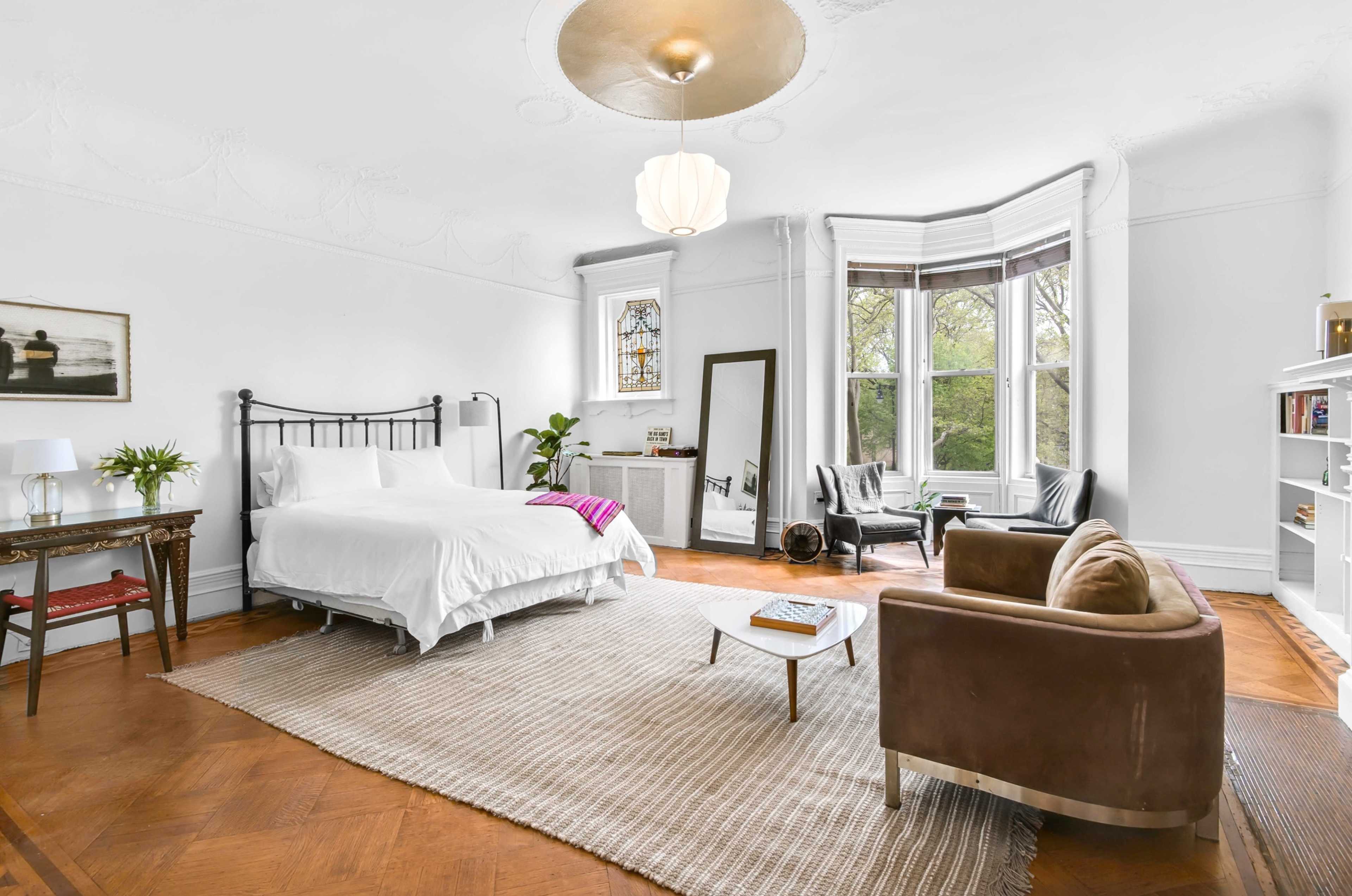 A spacious bedroom features a bed, a seating area with two chairs, a decorative mirror, and large windows allowing natural light to enter.