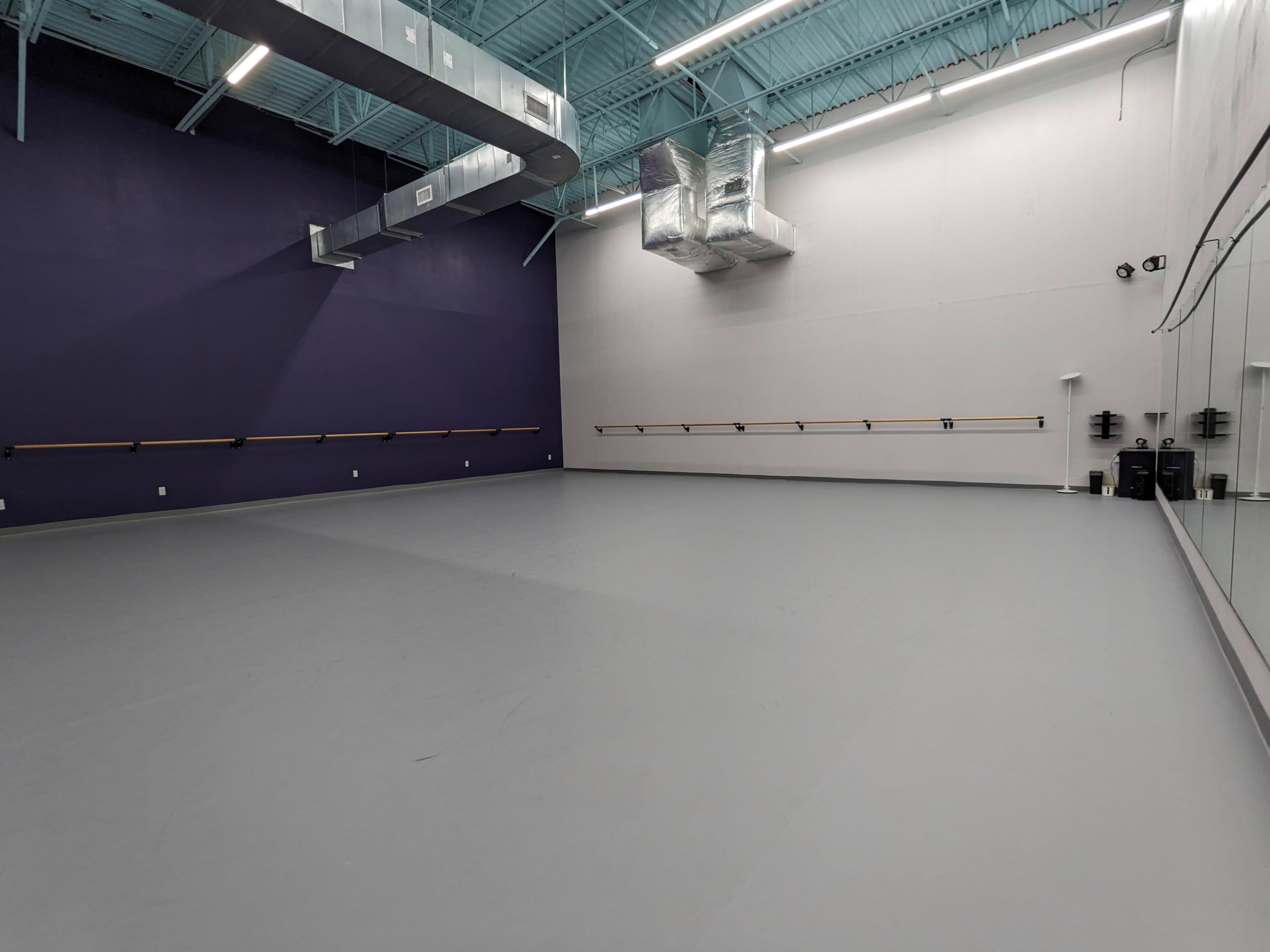 The image shows a spacious dance studio with a gray floor, purple walls, and mirrors along one side.