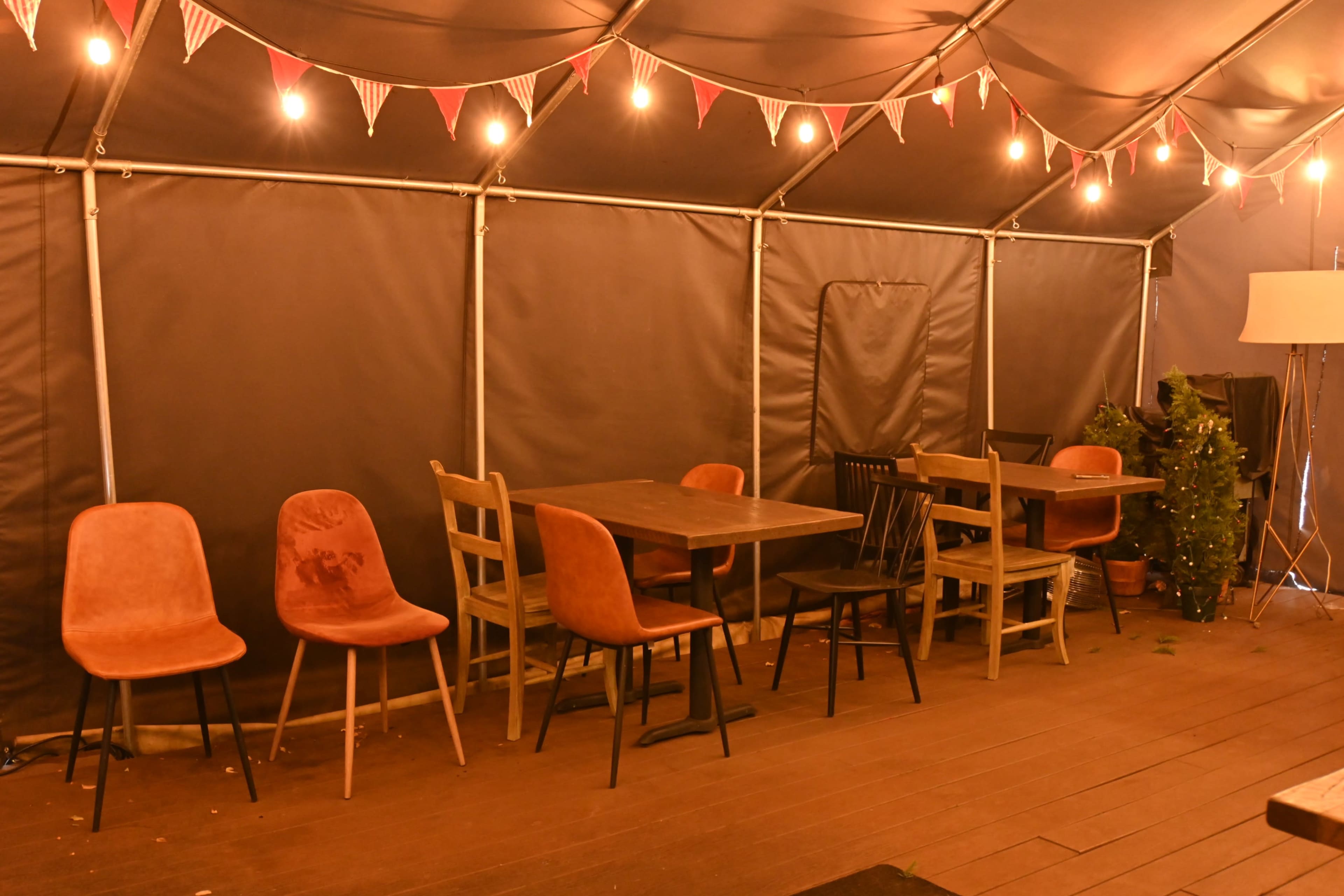 The image shows a tented dining area with several tables and chairs arranged, decorated with string lights and bunting.