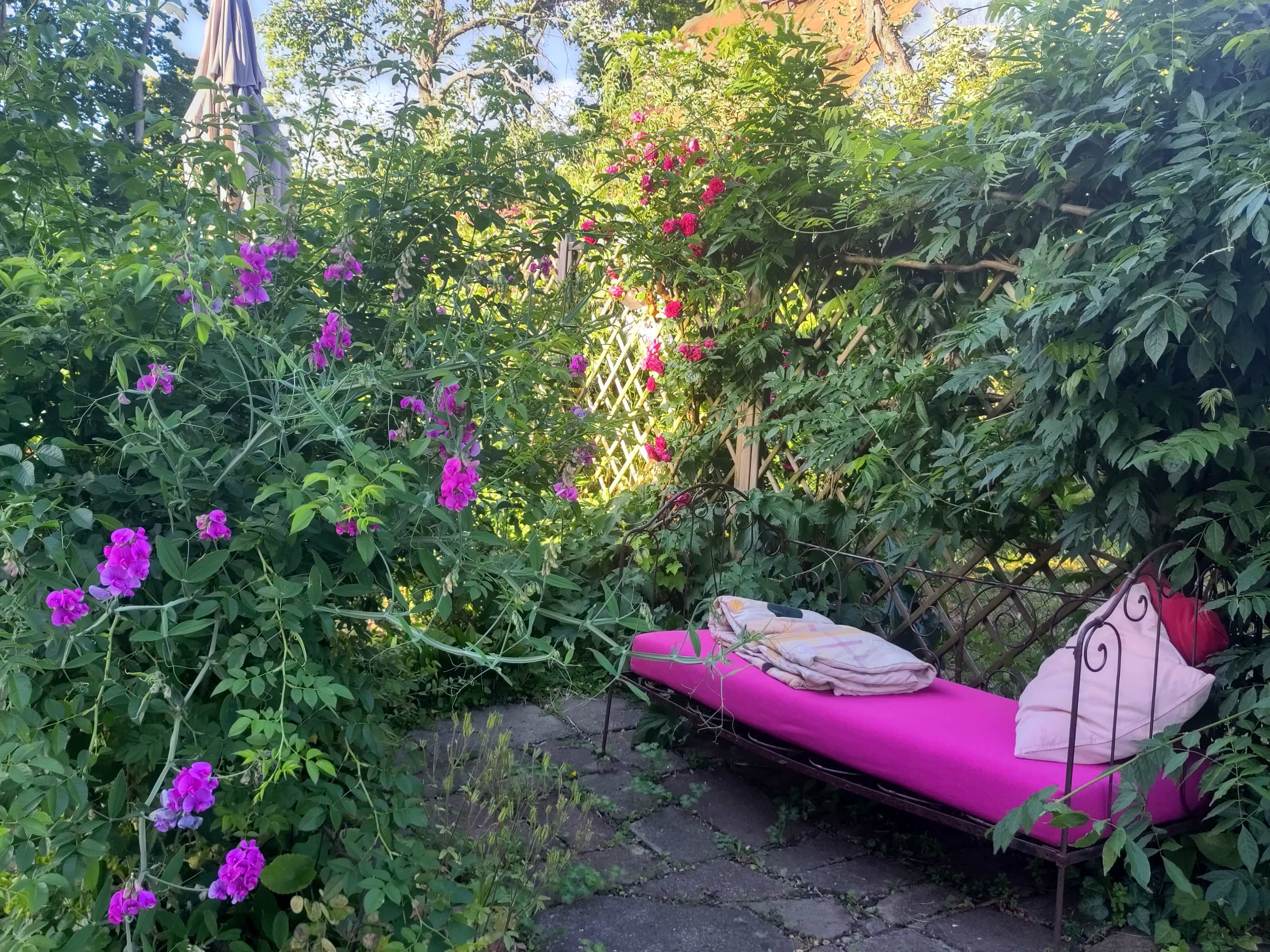 A daybed with pink cushions is nestled among lush greenery and vibrant flowers in a garden setting.