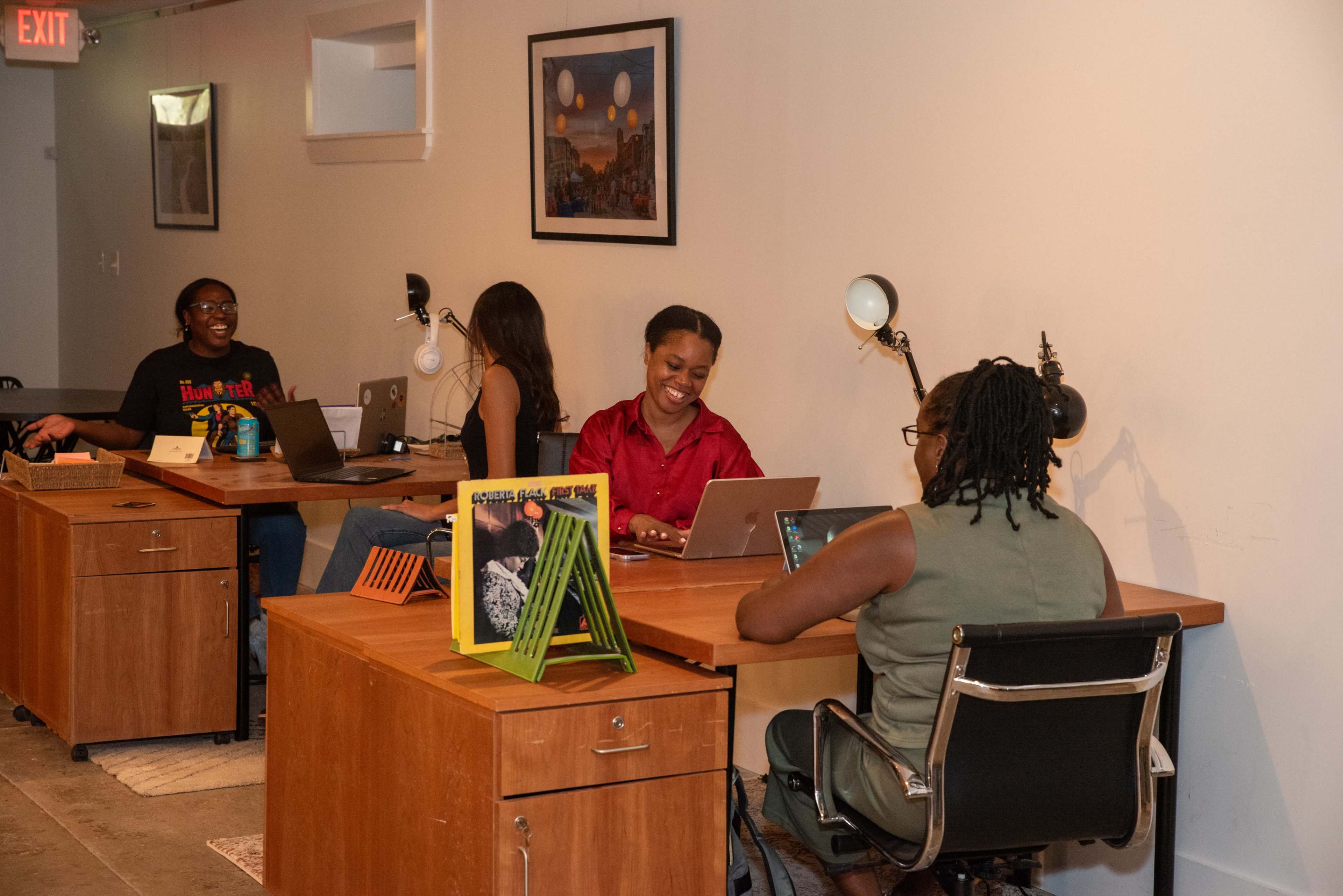 Four individuals are seated at wooden desks in a workspace, each engaged with their laptops, while a colorful book is displayed on one of the desks.