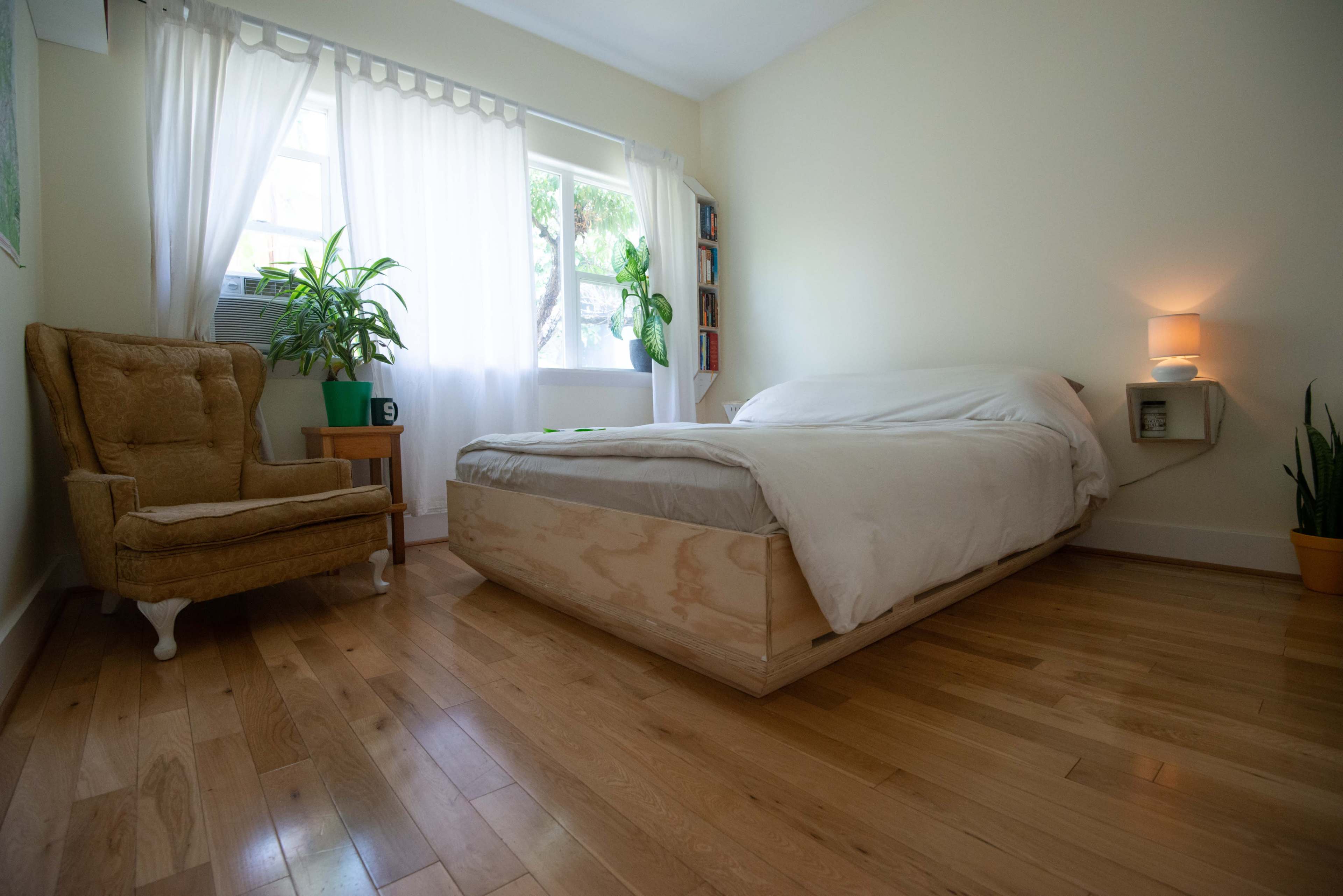 The room features a bed with a wooden platform, a chair, and several plants, all illuminated by natural light from a window.