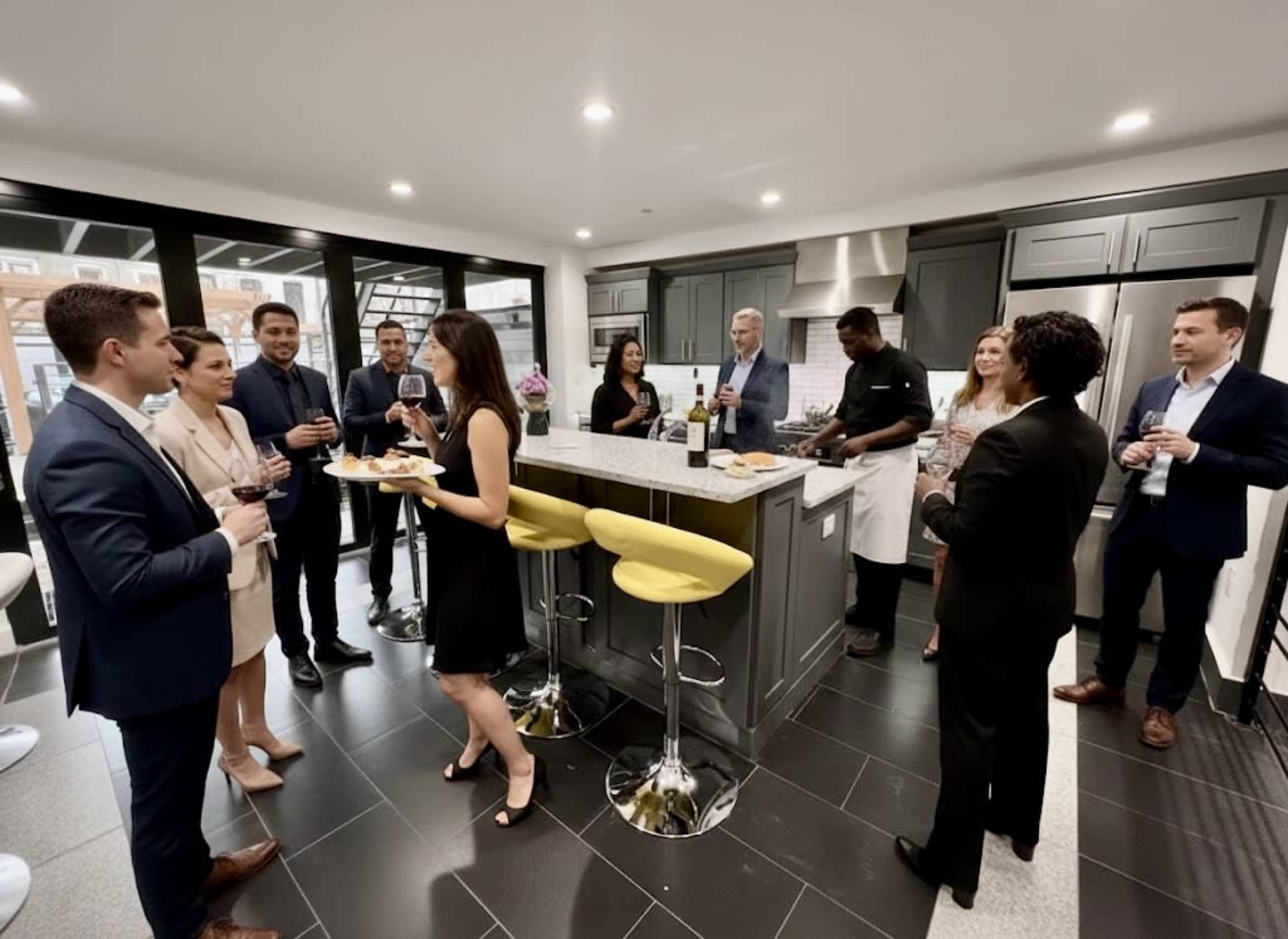 A group of professionals is socializing in a modern kitchen, with some holding drinks and food while others prepare in the background.
