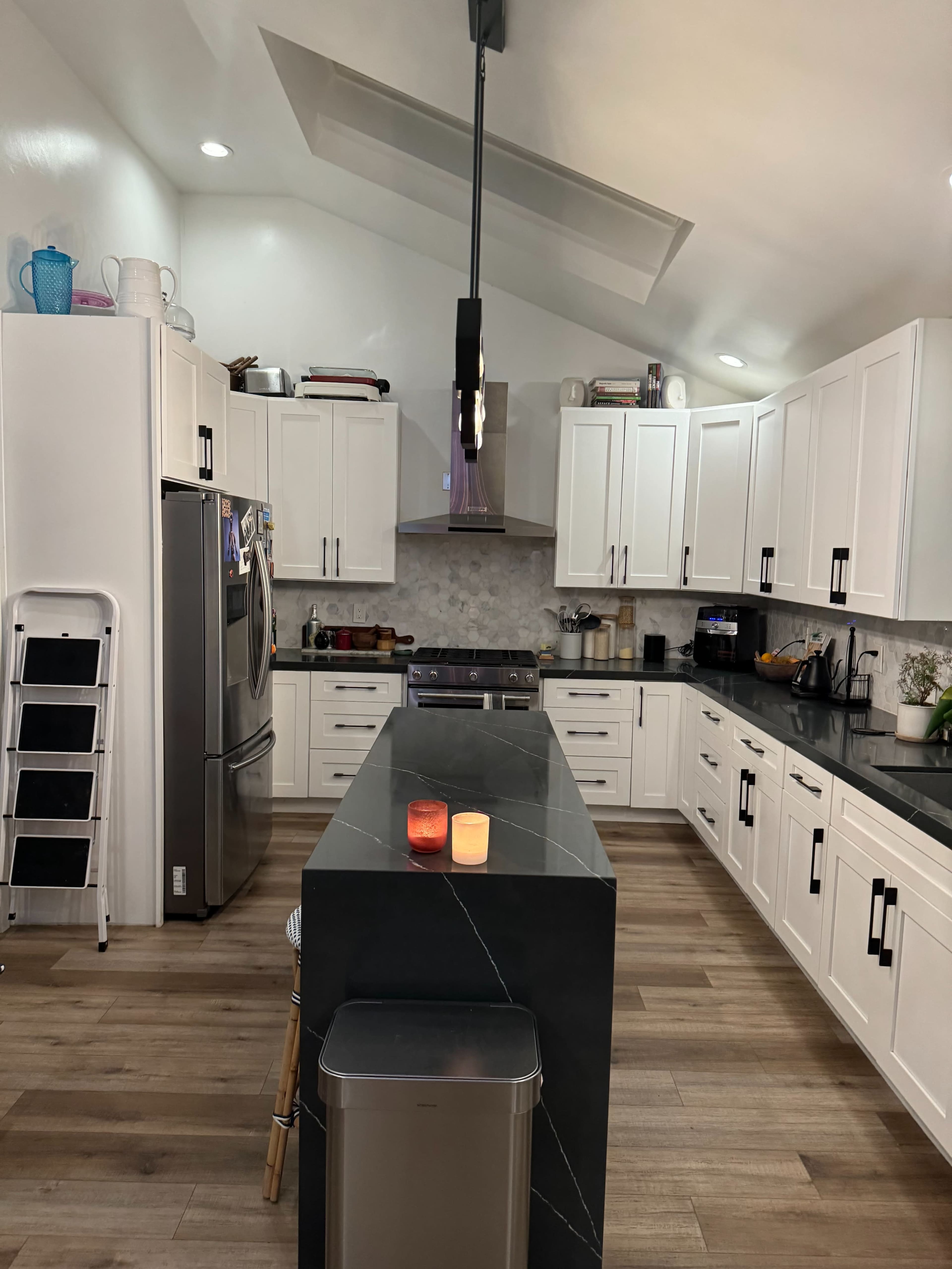 A modern kitchen features white cabinets, a large island with a black countertop, stainless steel appliances, and wooden flooring.
