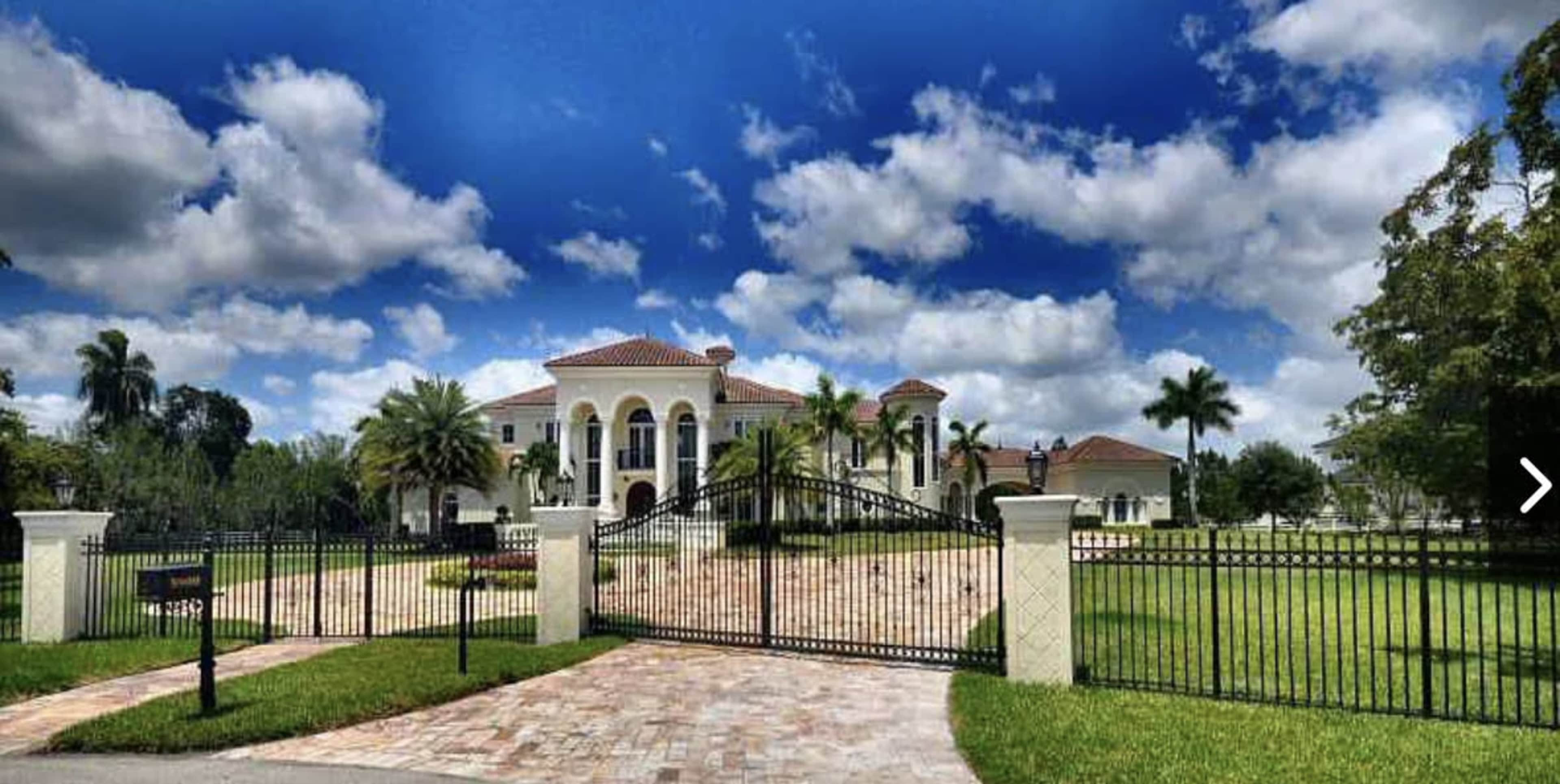 A large, two-story mansion with a red-tiled roof and palm trees is enclosed by a black wrought-iron gate, set against a backdrop of blue sky and scattered clouds.