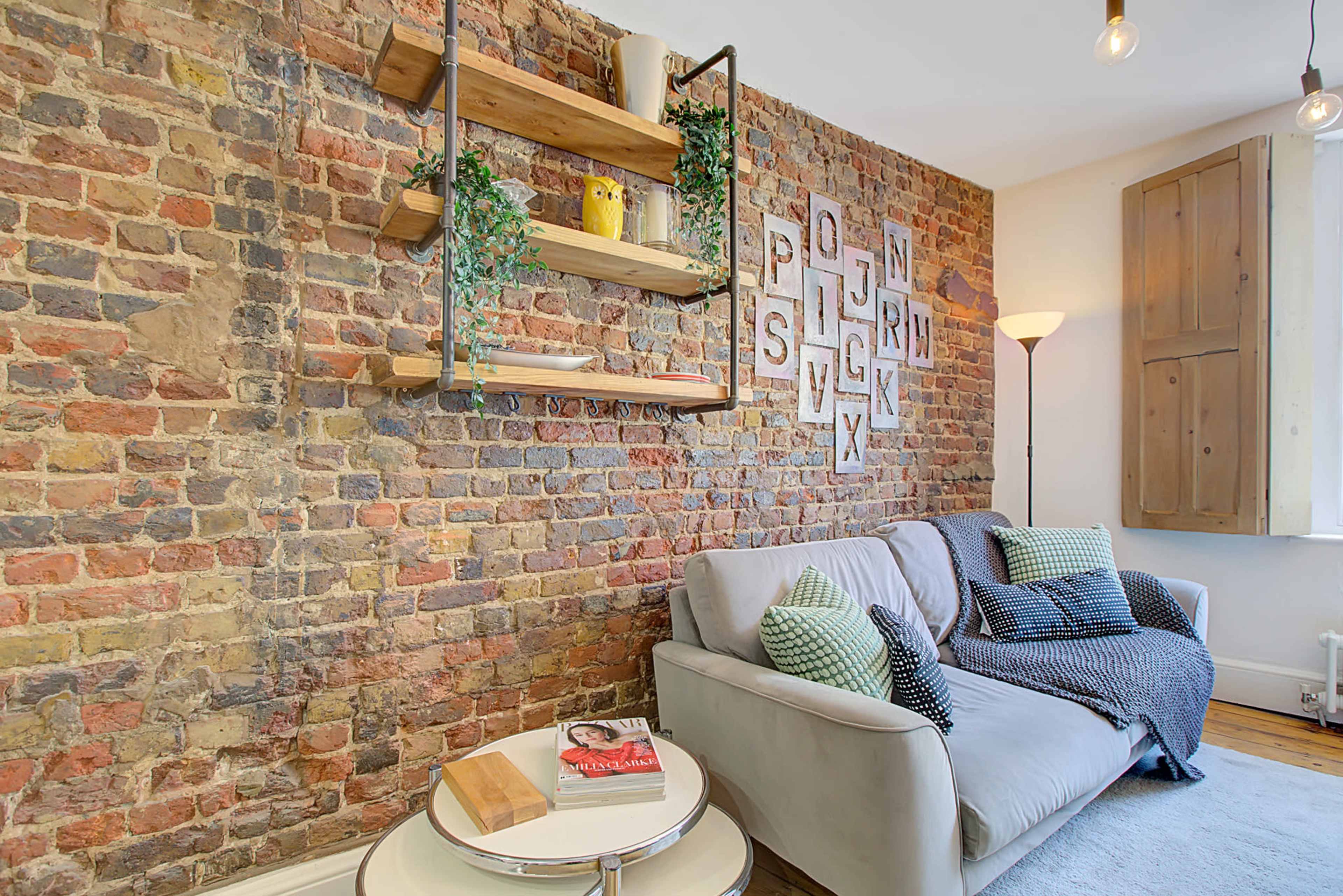 A cozy living room with a gray sofa, a round coffee table, and a brick wall featuring wooden shelves and decorative items.