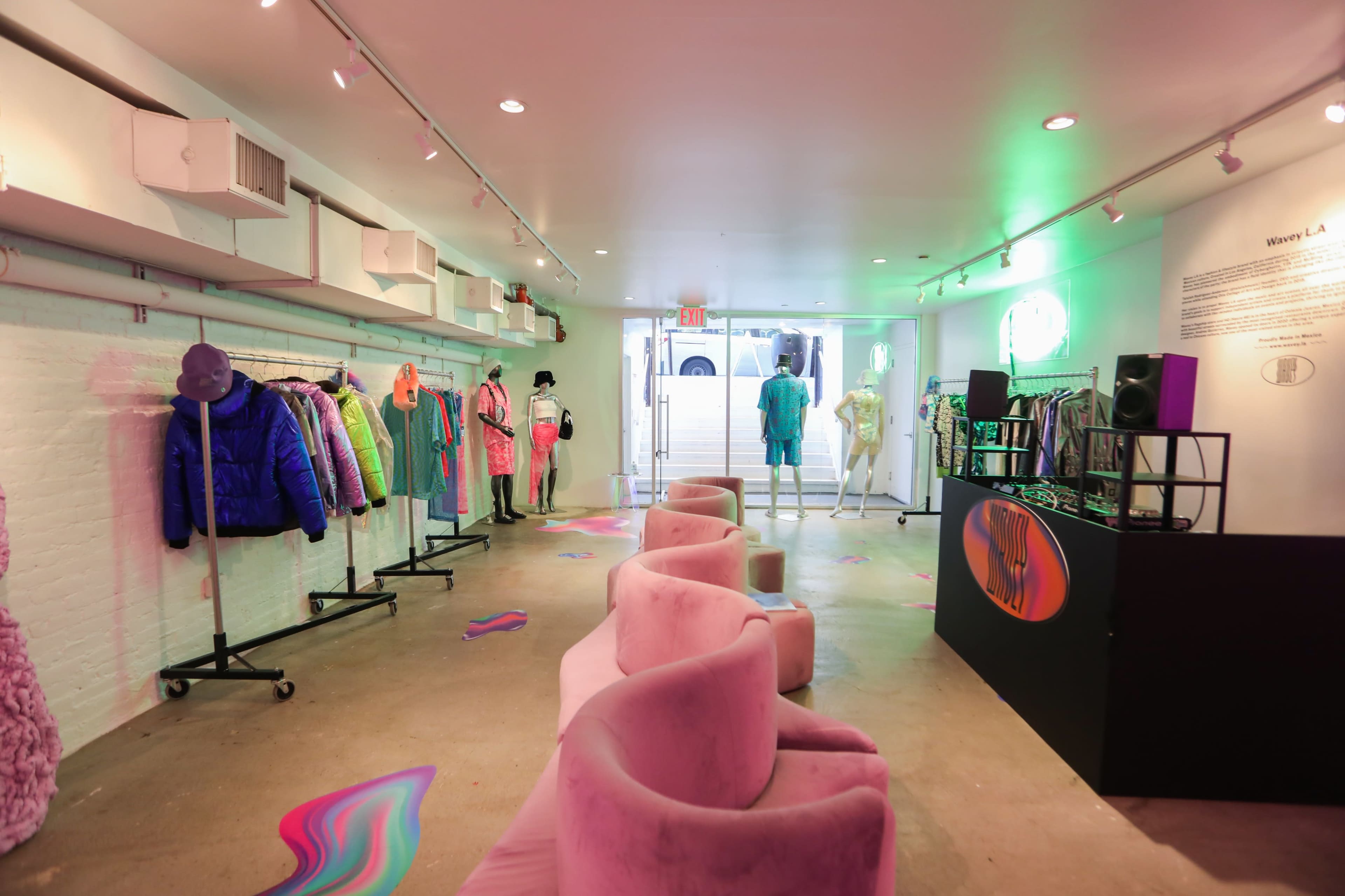 The interior of a modern clothing store features racks of colorful apparel on one side and a seating area with curved pink chairs on the other, with a DJ setup visible near the entrance.