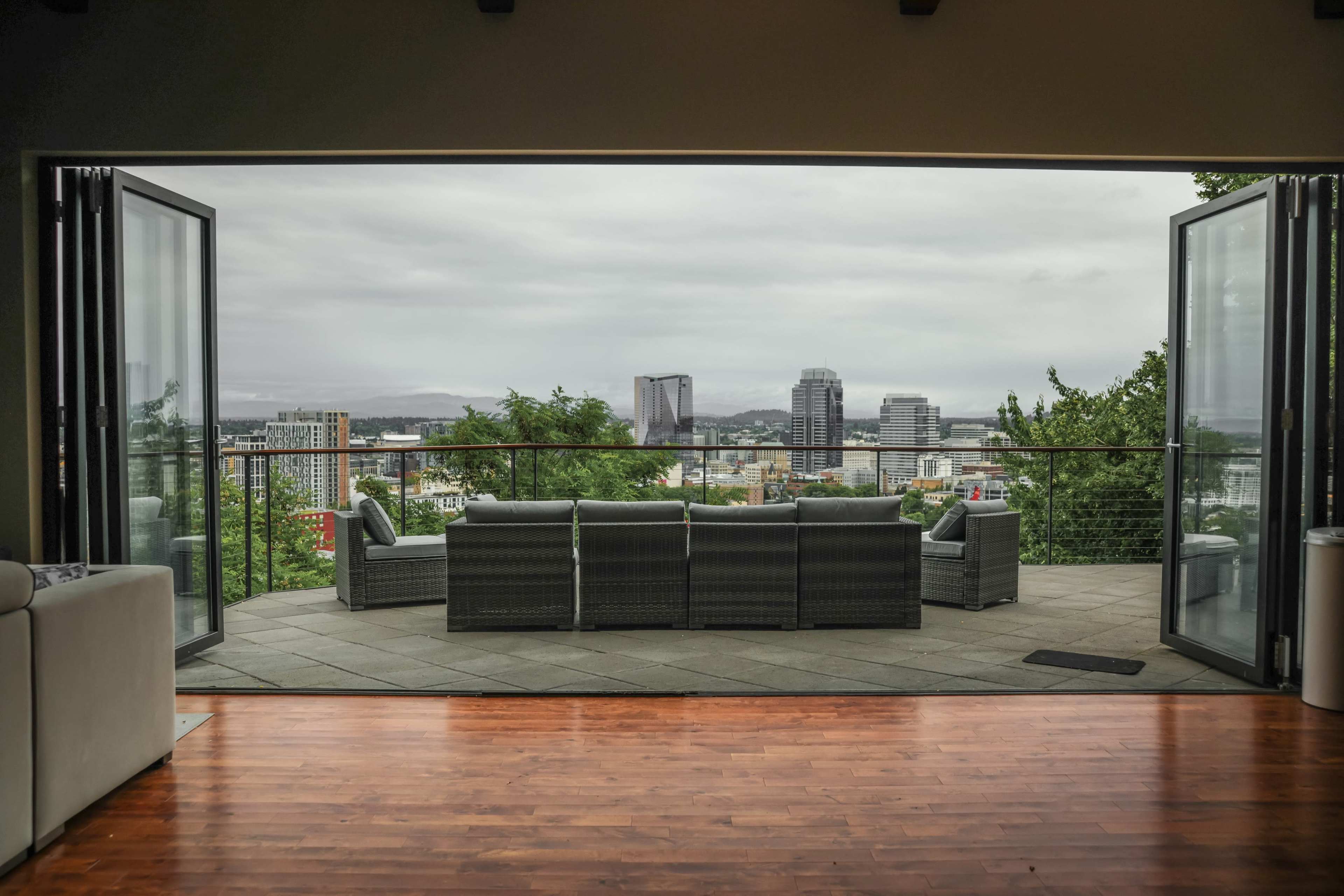 A spacious patio with outdoor seating opens up to a panoramic view of a cityscape under cloudy skies.