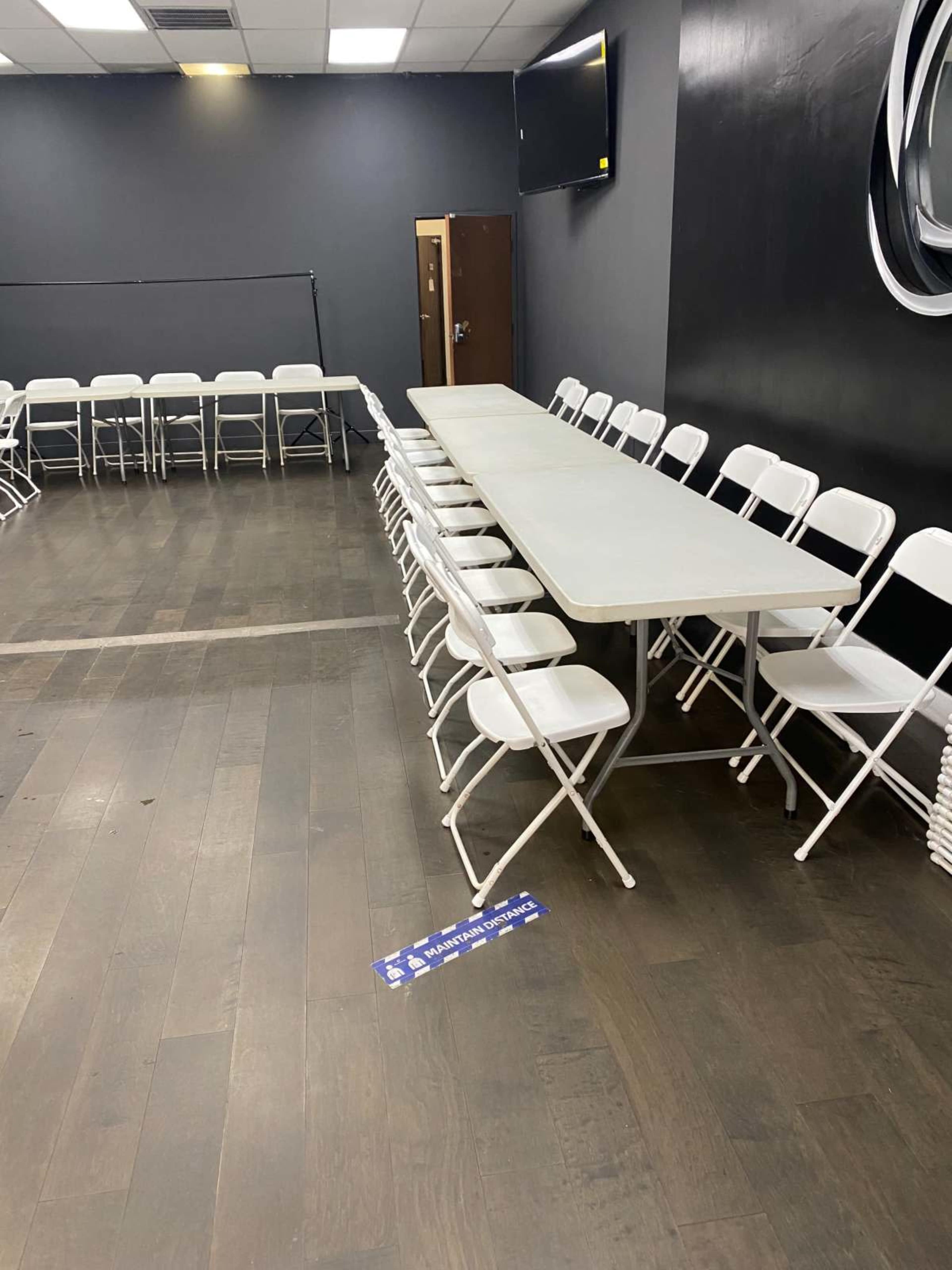 A long table is set up in a room with white folding chairs arranged on either side, and a television mounted on the wall.