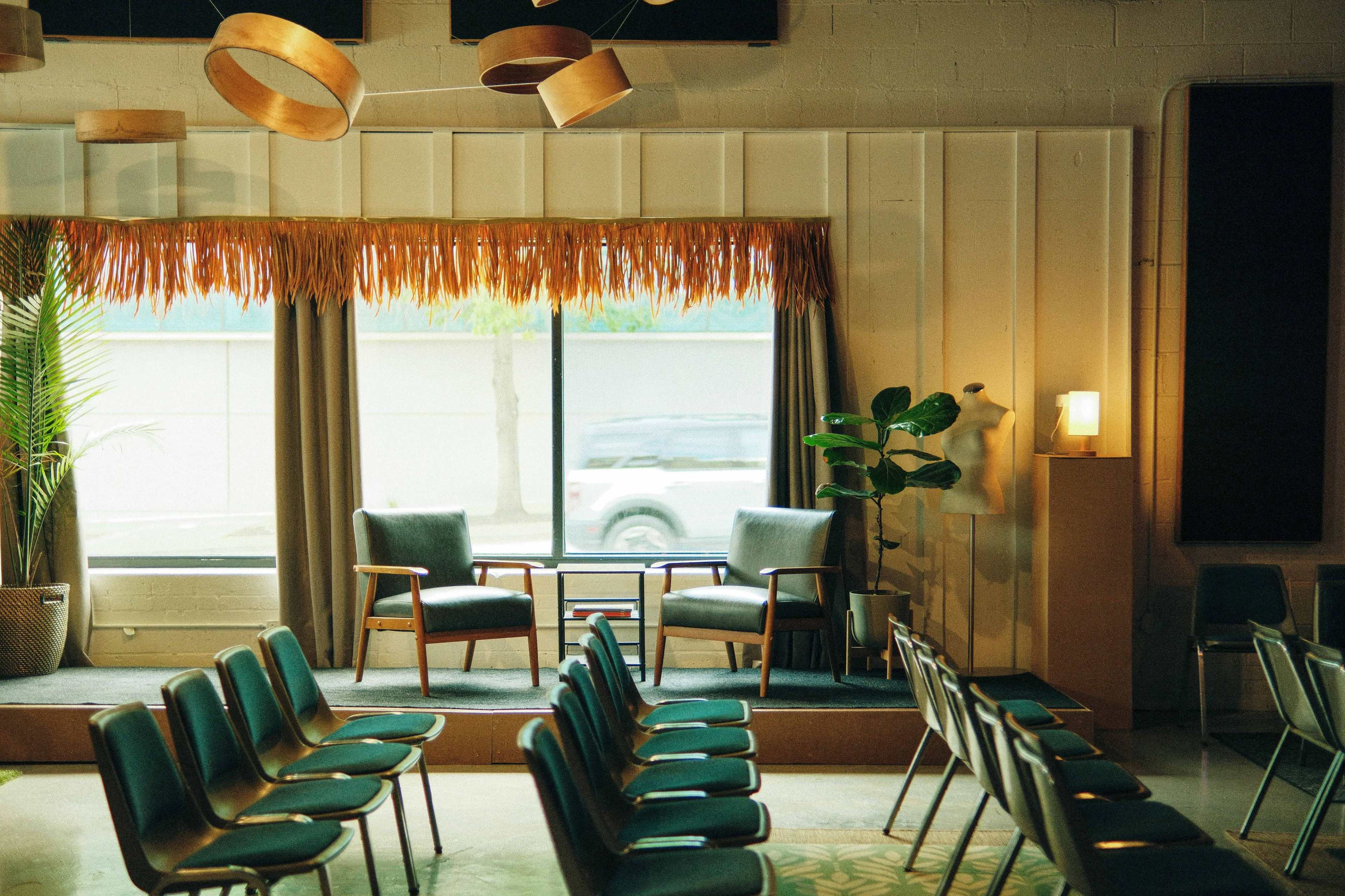 A seated area with two chairs and a small table is framed by a large window and decorative curtains, while rows of empty chairs face the stage.