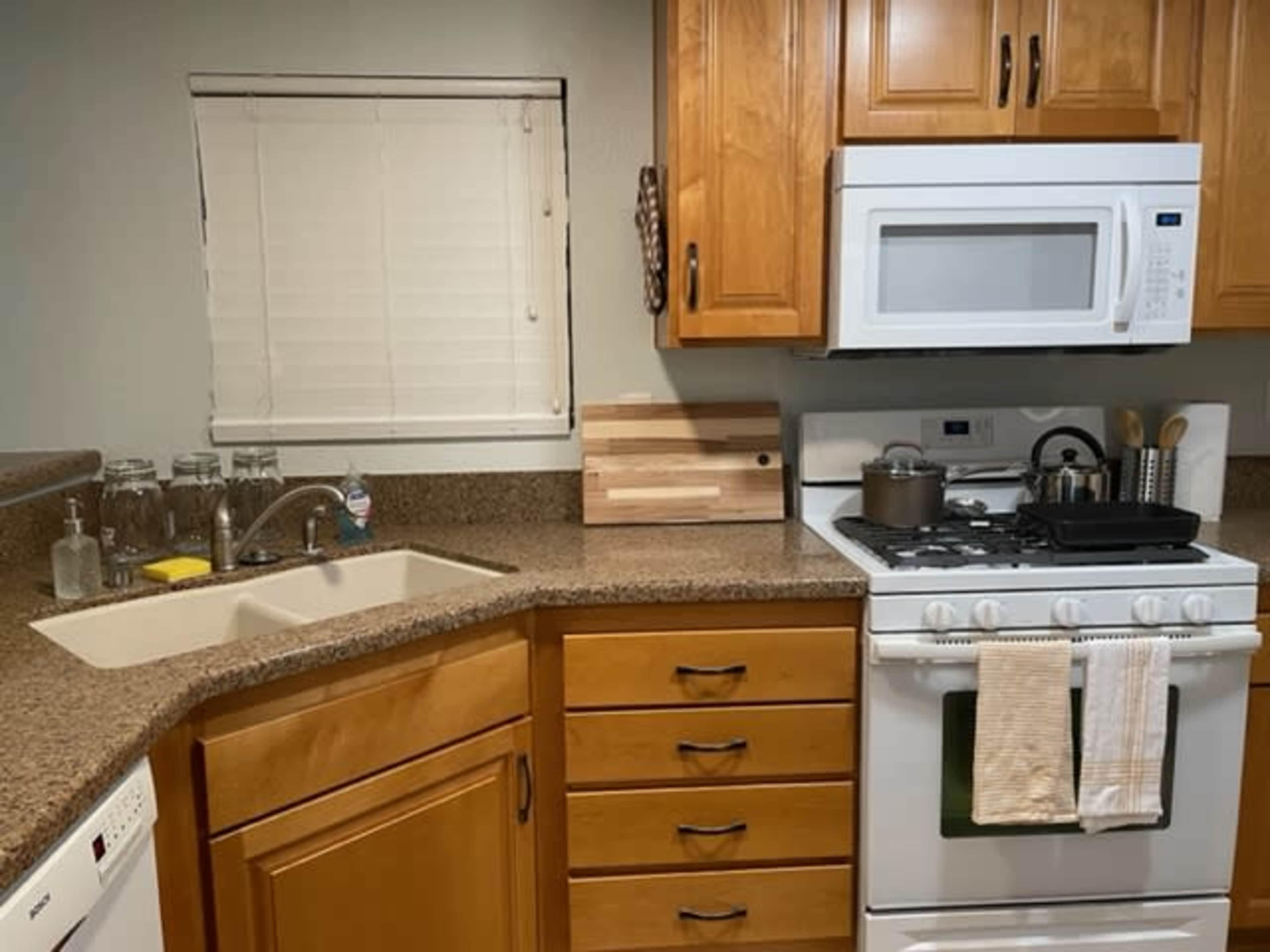 The image shows a kitchen with wooden cabinets, a double sink, a stove with a pot, and jars on the counter beside a window.