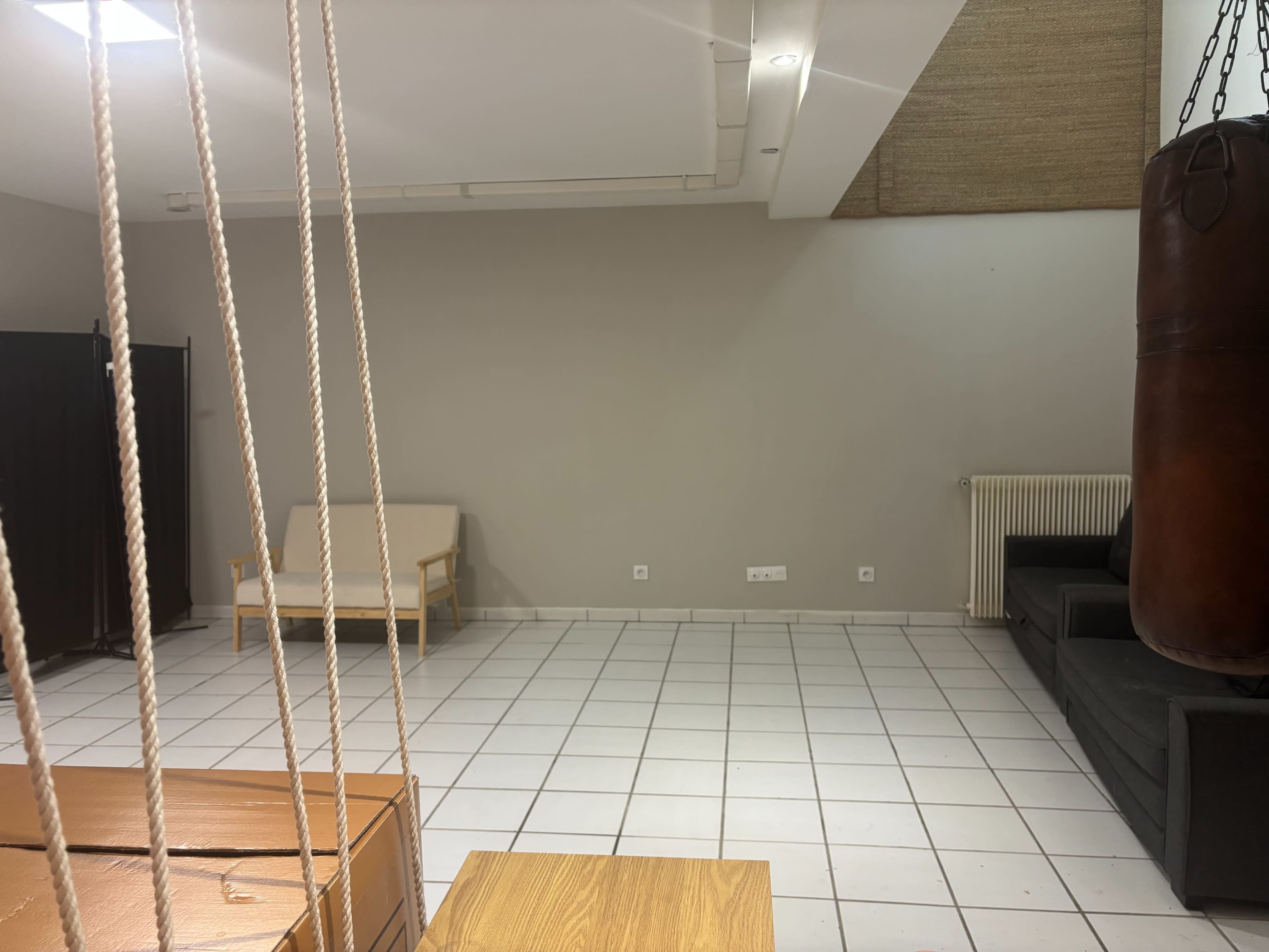 The image depicts a spacious room featuring a punching bag, a wooden table, a couch, and tiled flooring.