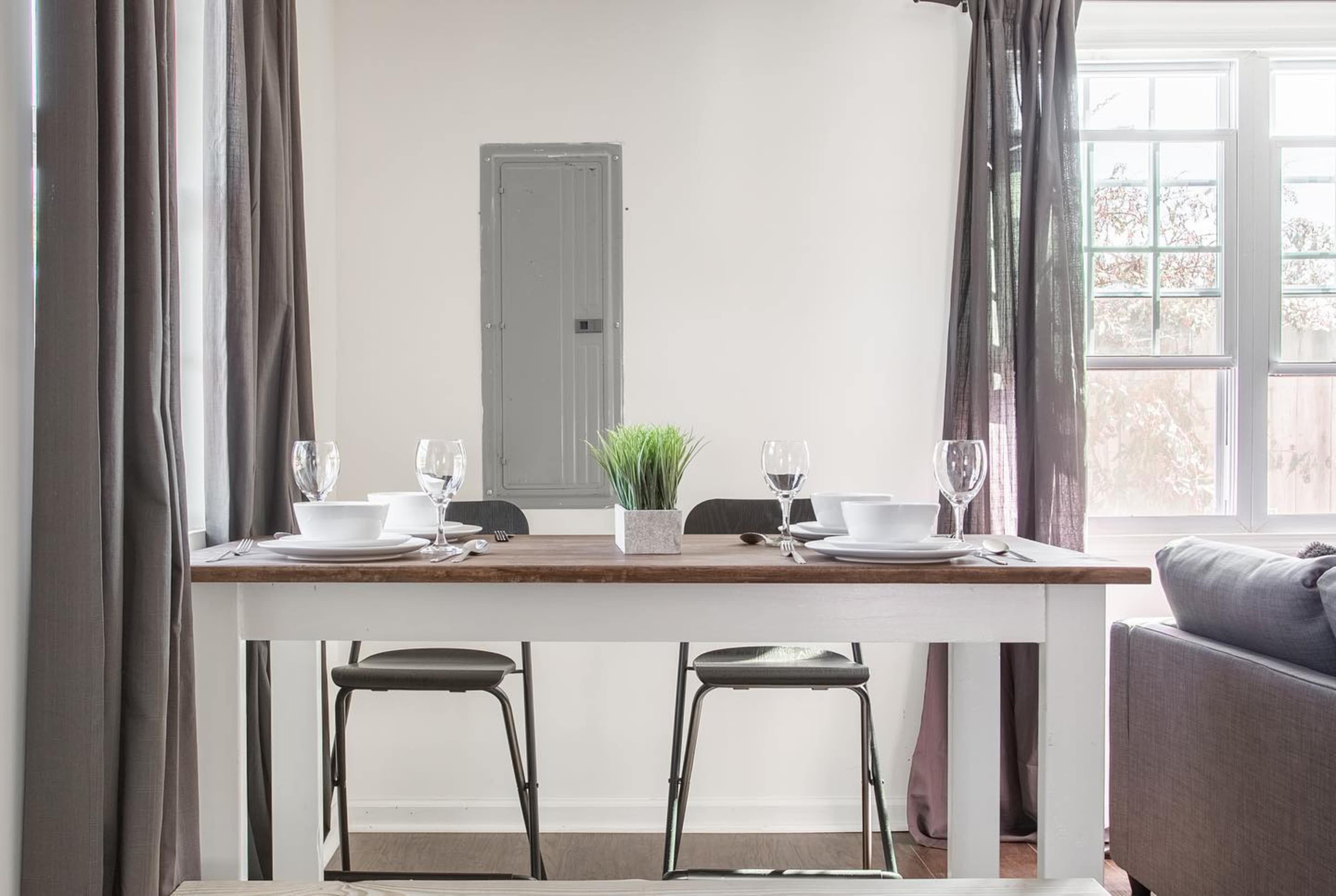 A dining table set for four with glassware and plates is positioned in a bright room with curtains and a small window.