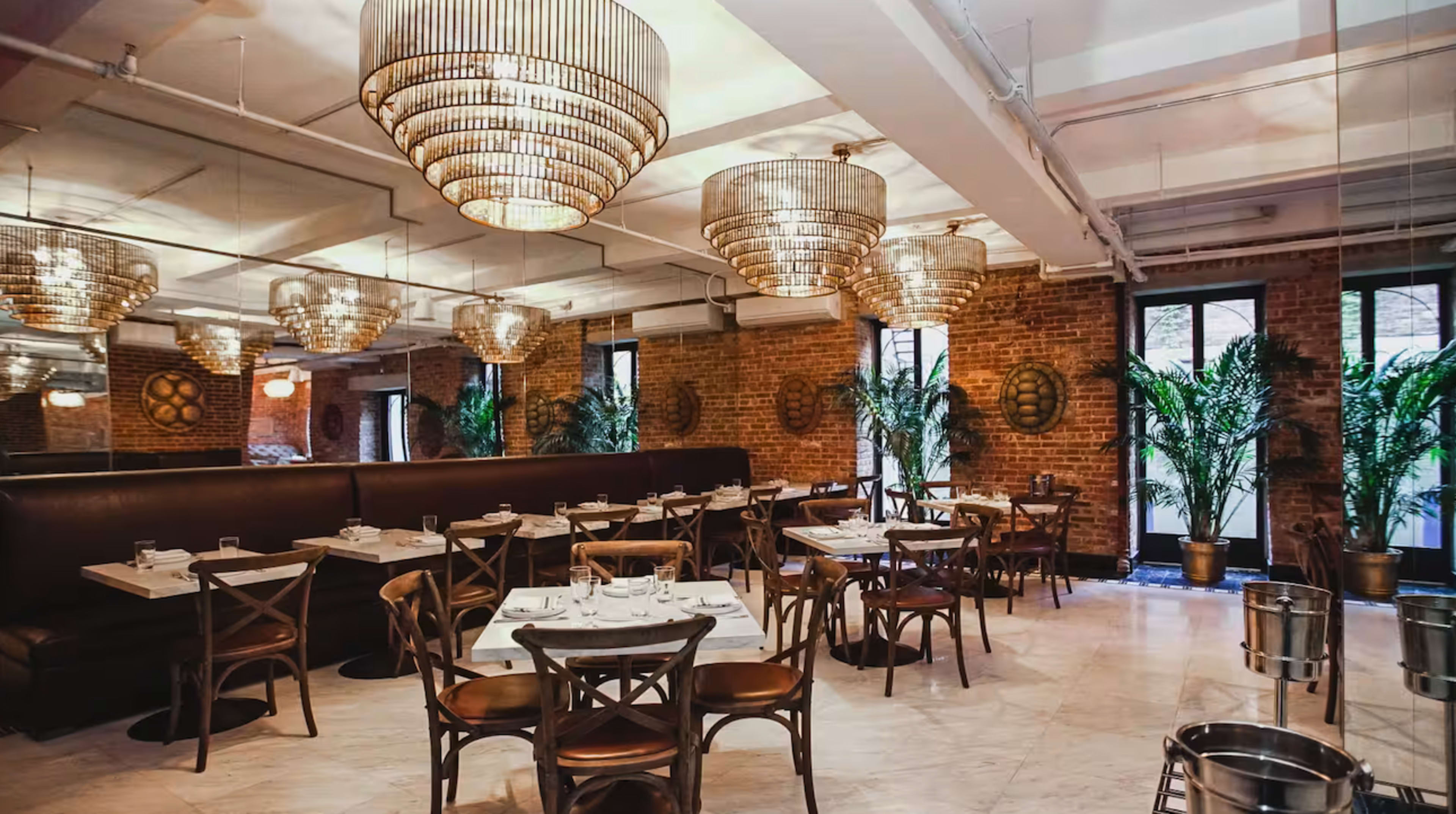 The image shows a modern restaurant interior featuring marble tables, wooden chairs, and large chandeliers, complemented by greenery and exposed brick walls.
