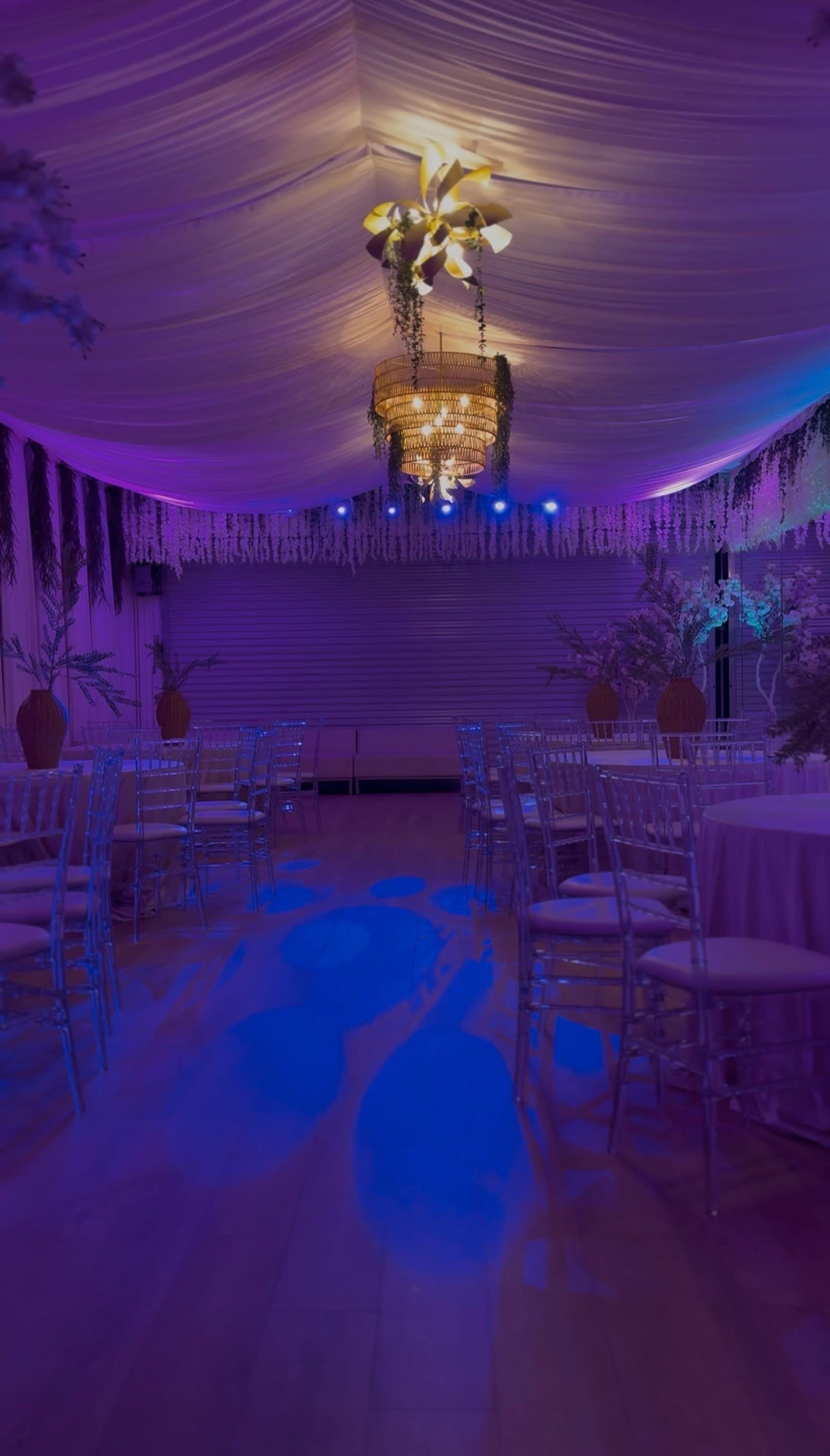A decorated indoor venue features round tables with white tablecloths, silver chairs, and colorful lighting illuminating the ceiling and floor.