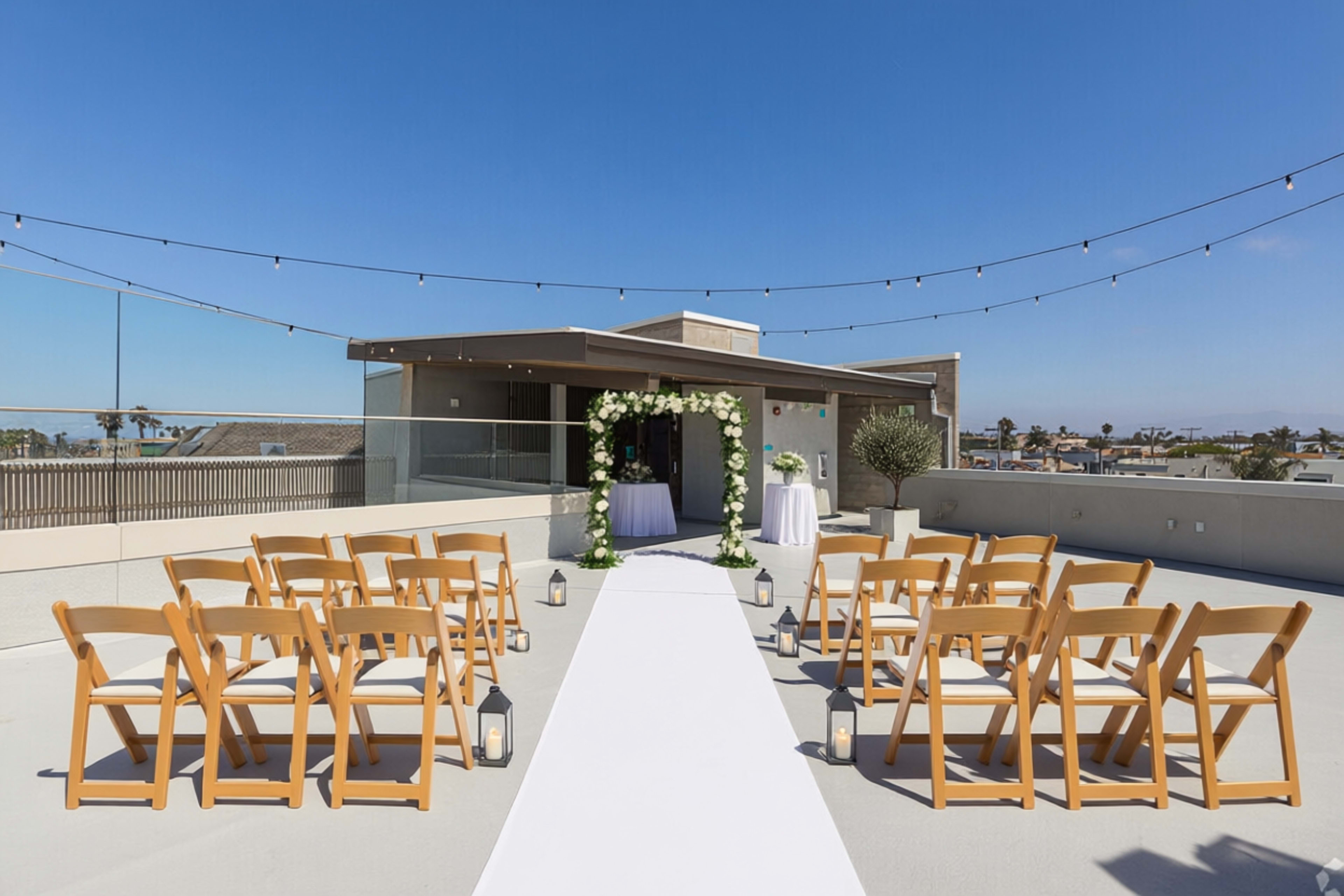 A rooftop wedding venue features a white aisle runner leading to an arch adorned with flowers, surrounded by wooden chairs and string lights overhead.