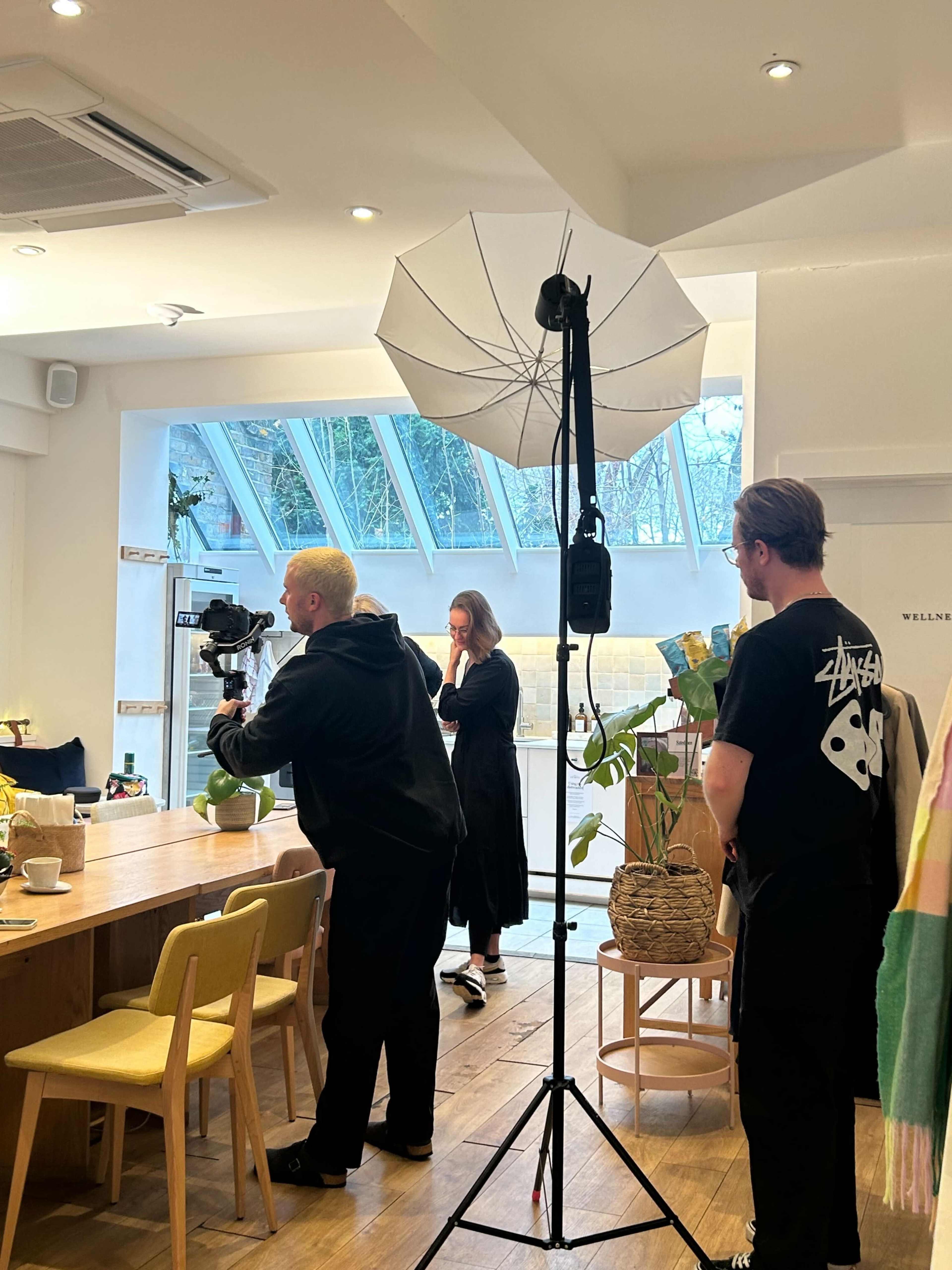 A camera crew is set up in a bright café, with one person filming while another adjusts lighting equipment as a third person observes.