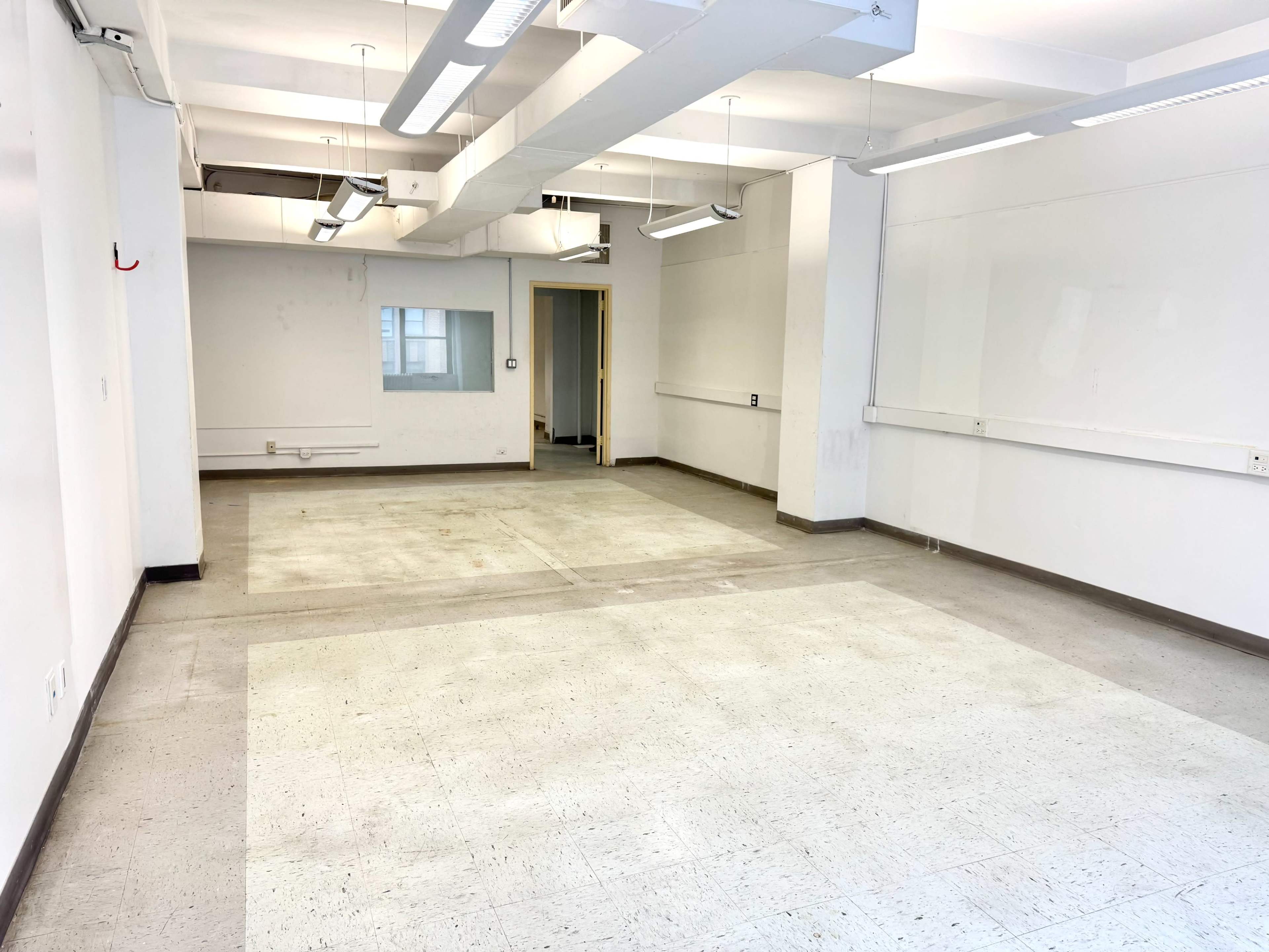 The image shows a vacant room with white walls, a few fluorescent lights, and an unfinished floor.