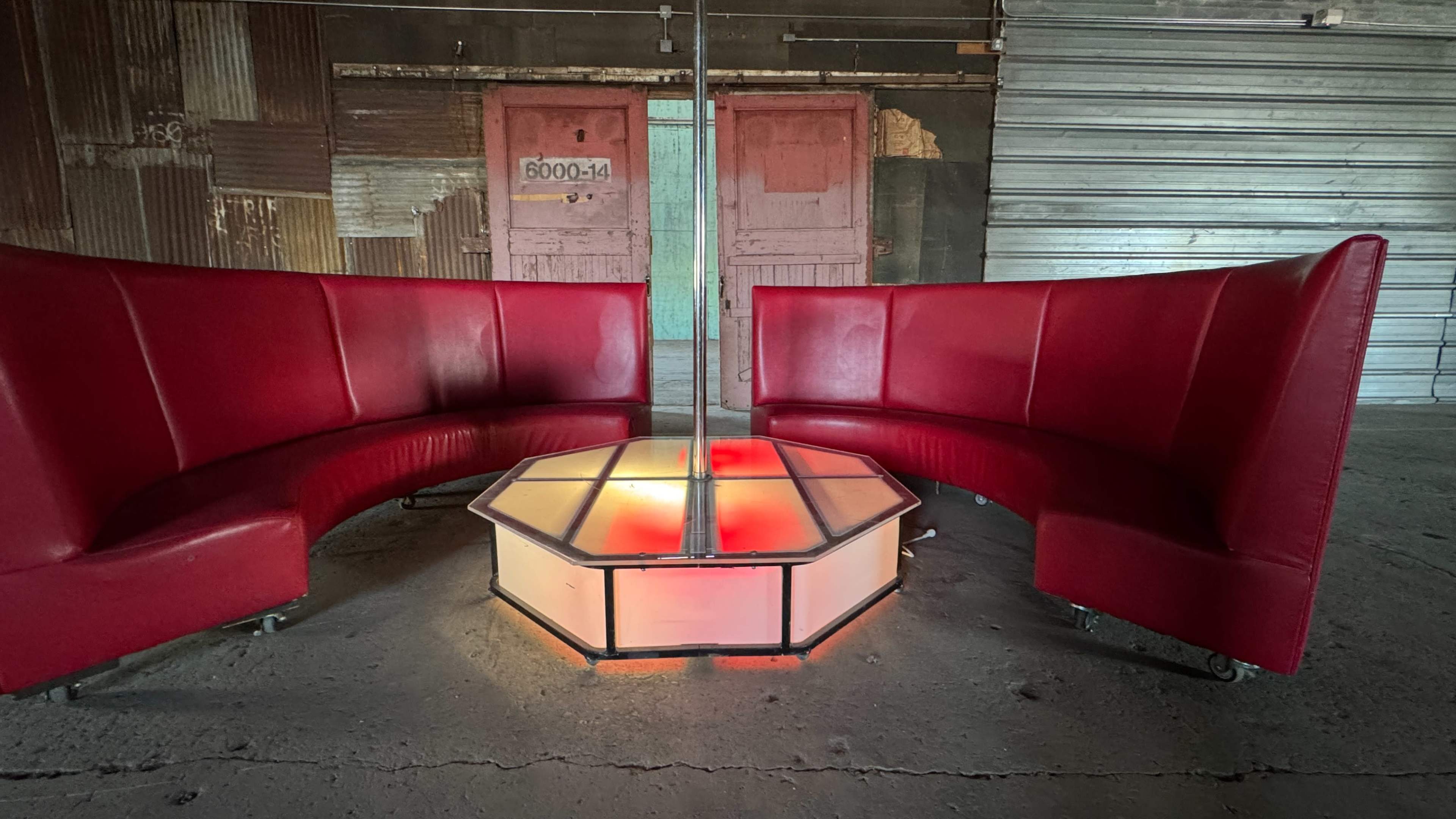 The image depicts a red booth seating arrangement surrounding a central illuminated octagonal platform in an industrial space with metal doors in the background.