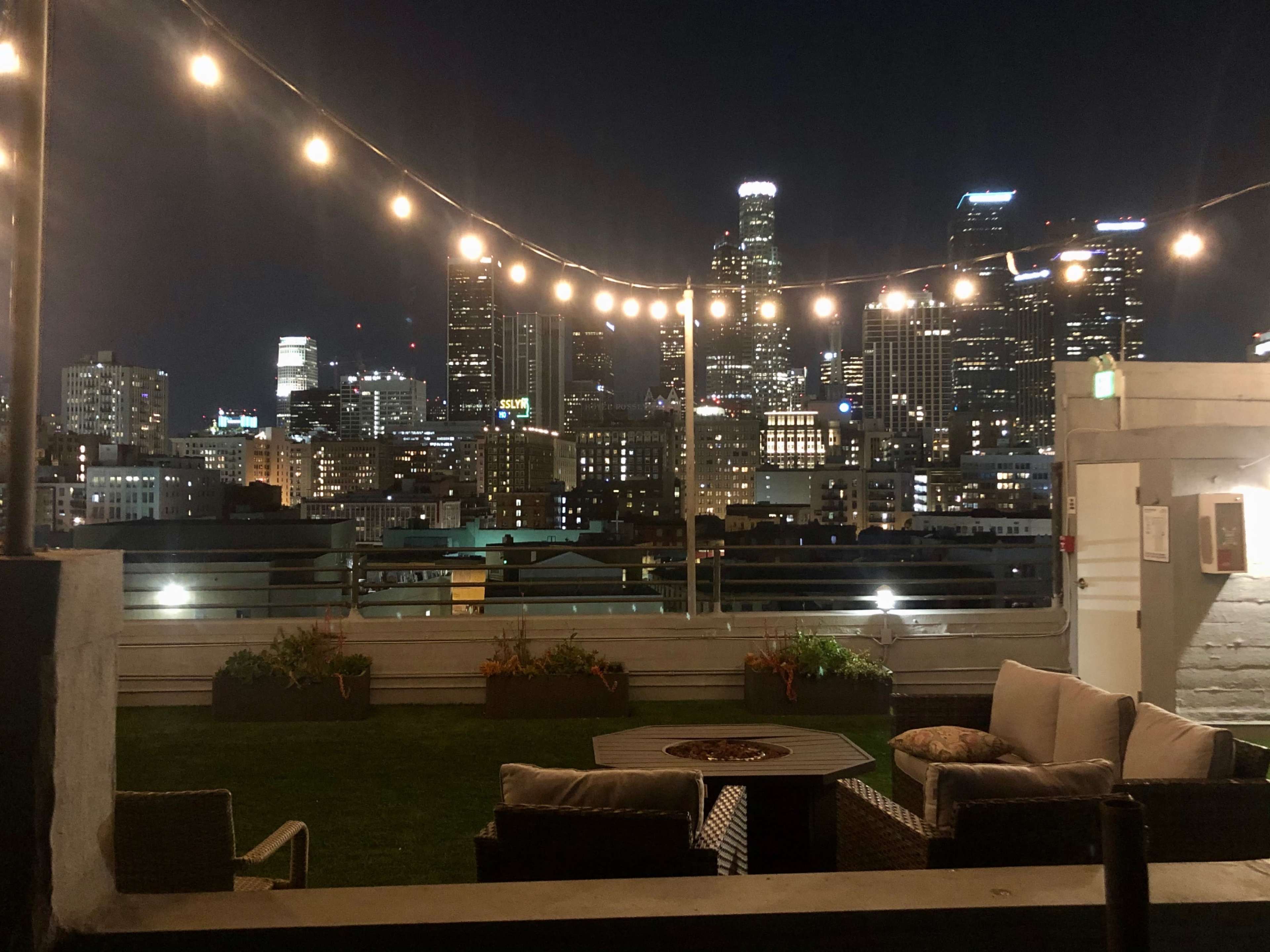 A rooftop lounge area with string lights overhead, overlooking a city skyline at night.