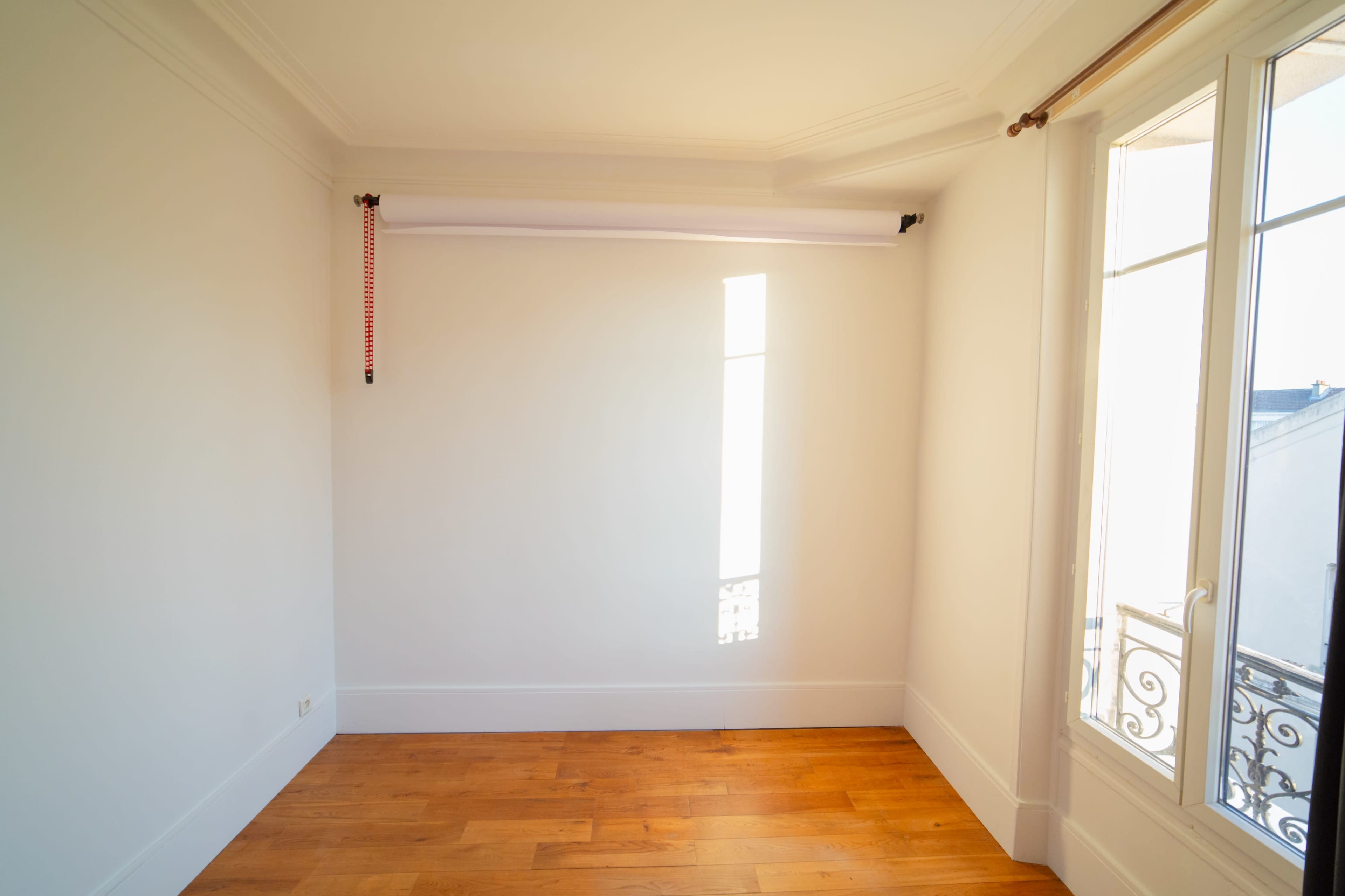 A bright, empty room features a wooden floor and a large window with a decorative balcony railing, allowing natural light to enter.
