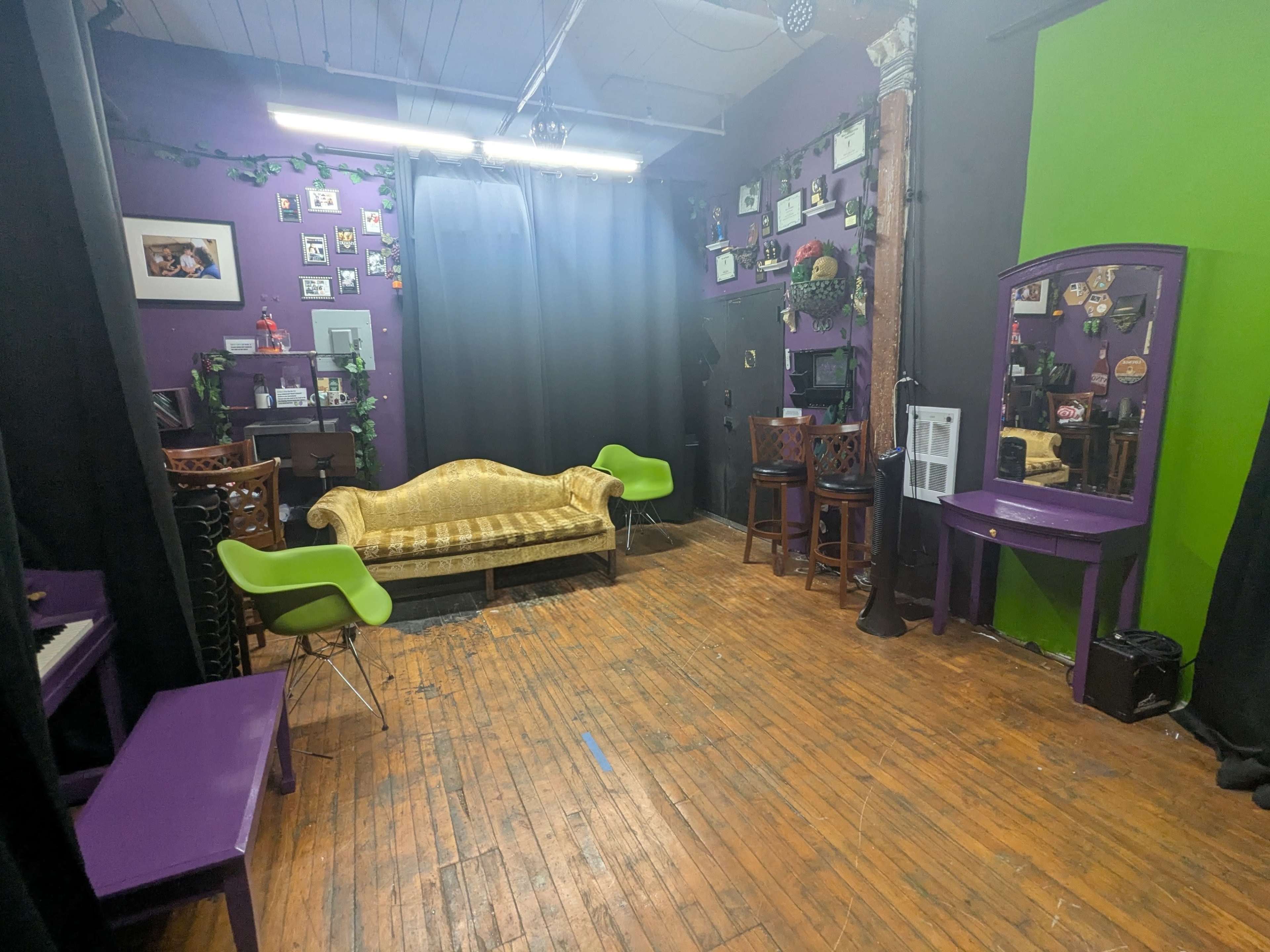 The image shows a room with purple and green walls, featuring a golden couch, several wooden bar stools, and a mirrored vanity against one wall.