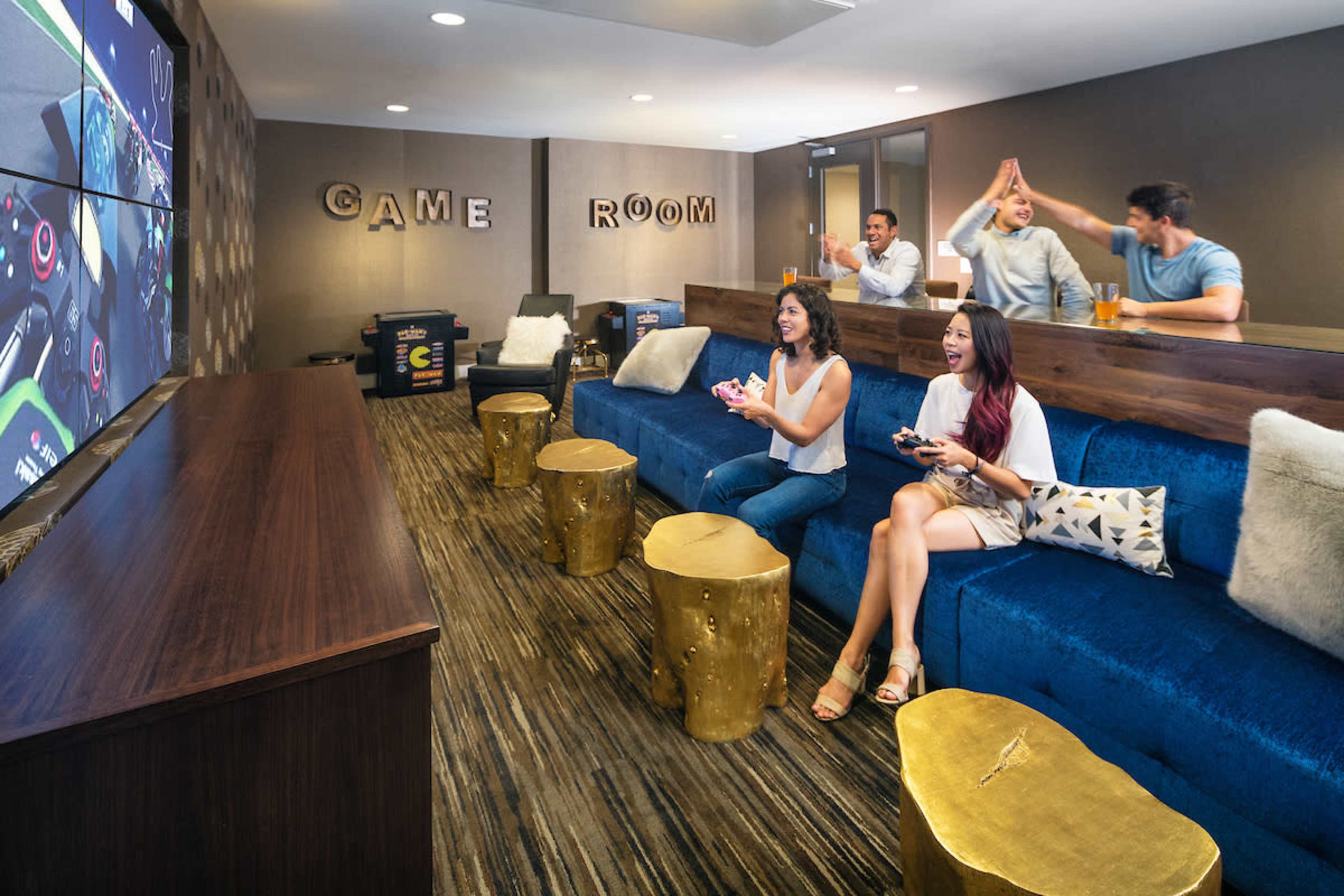 A group of five people enjoys playing video games in a game room equipped with large screens and seating.