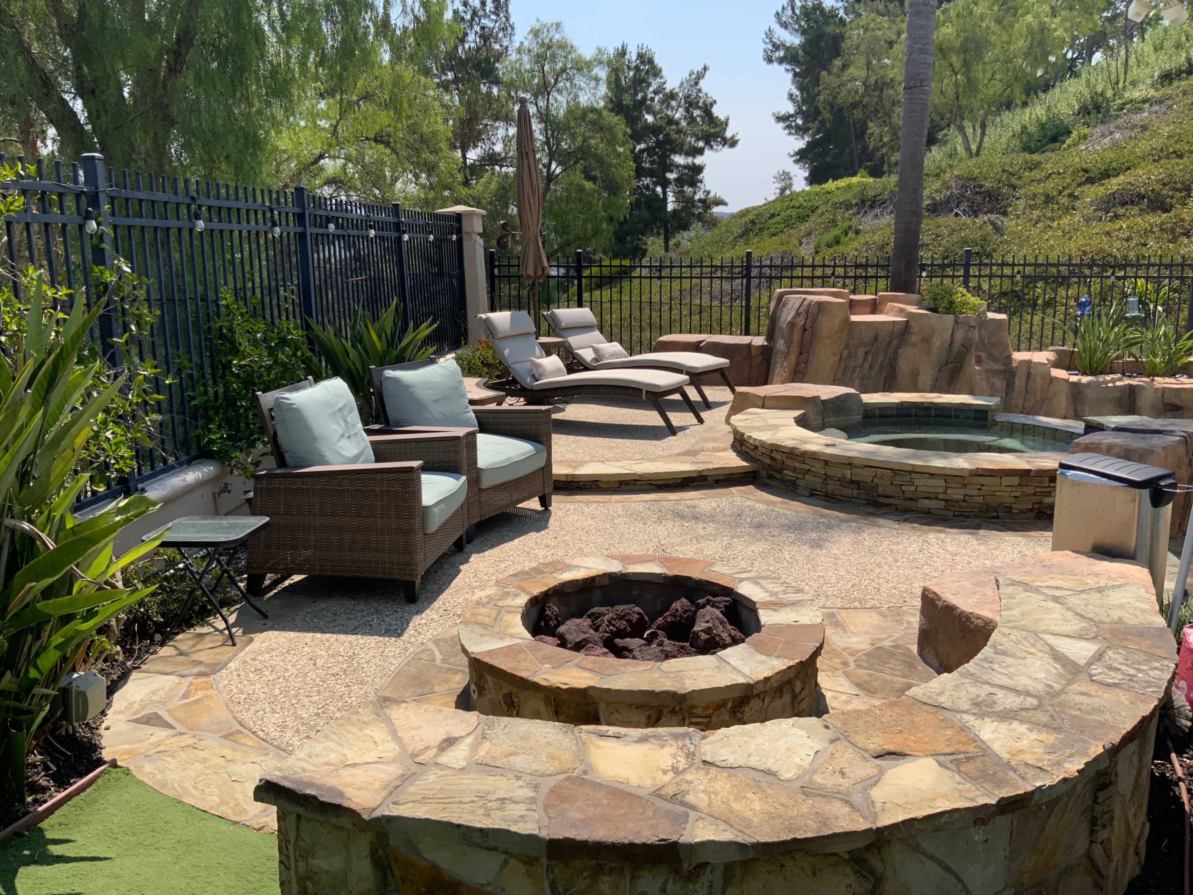 The image shows a landscaped backyard featuring a stone fire pit, a circular seating area, lounge chairs, and a hot tub surrounded by greenery.