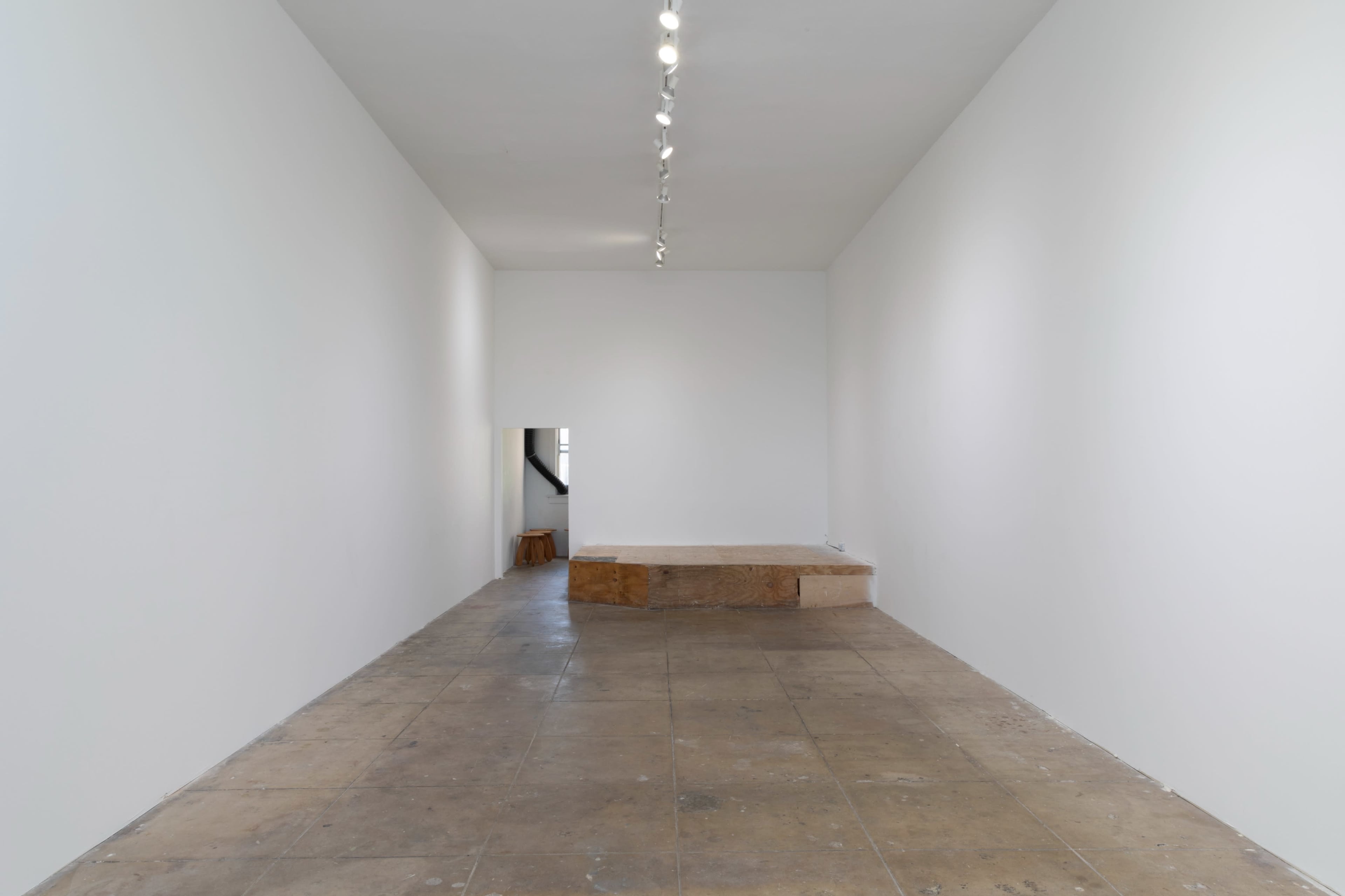 A minimalist art gallery featuring a stark white space with a wooden platform positioned against one wall.