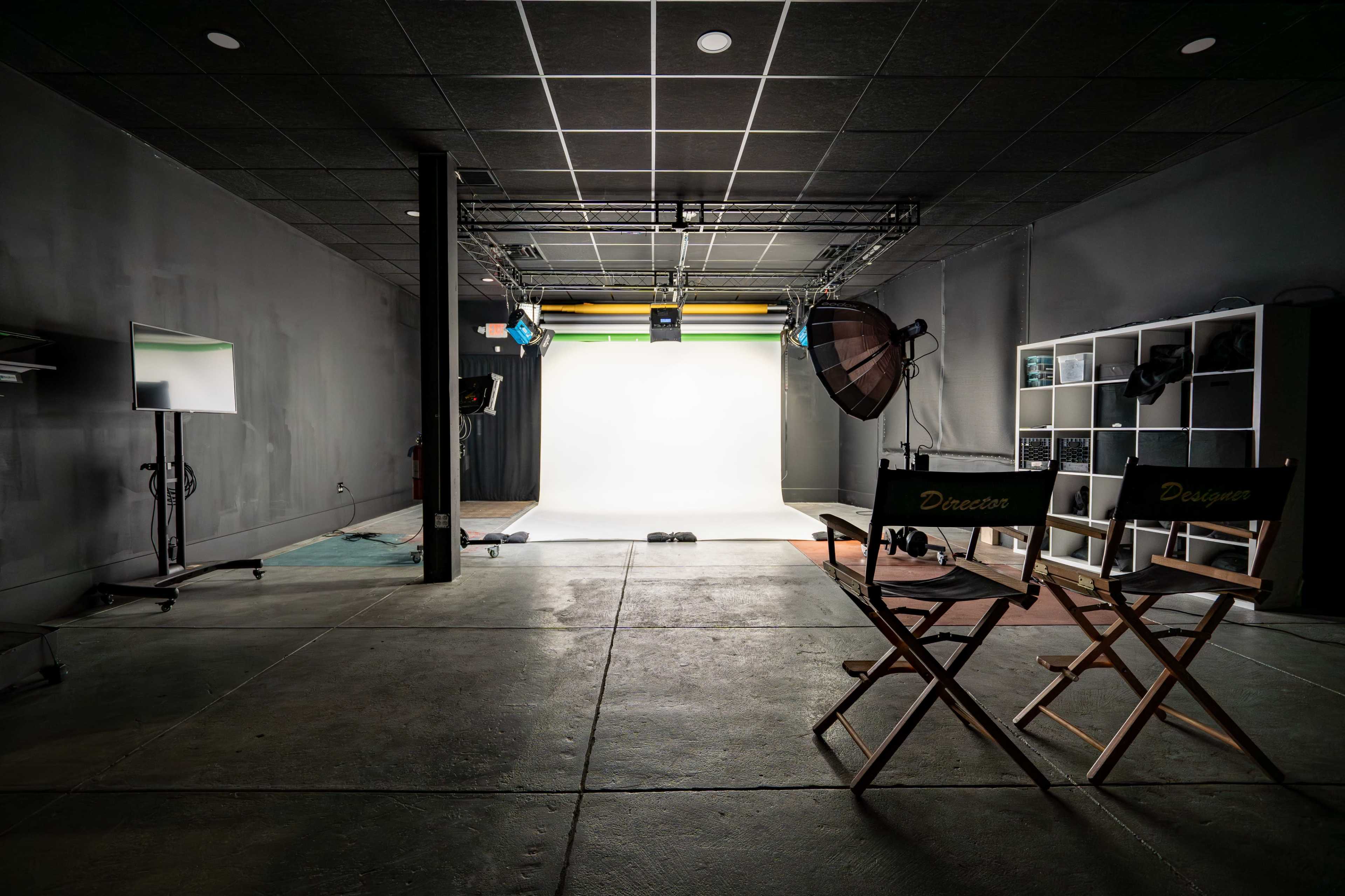 The image shows an empty photography studio with a white backdrop, lighting equipment, and director's chairs labeled "Director" and "Designer."