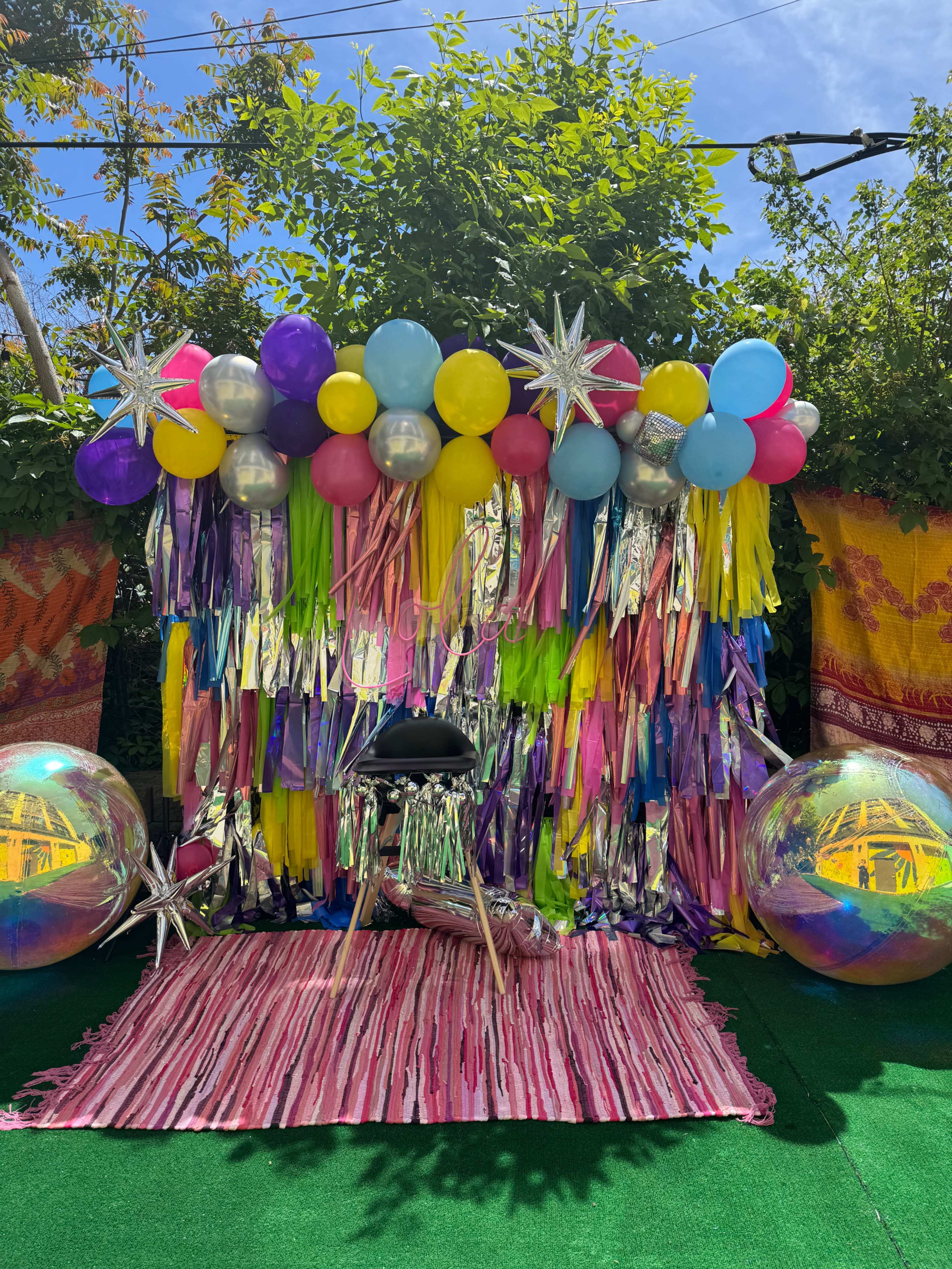 The image features a colorful party backdrop decorated with balloons, shiny streamers, and large reflective spheres, set on a vibrant pink and striped rug.