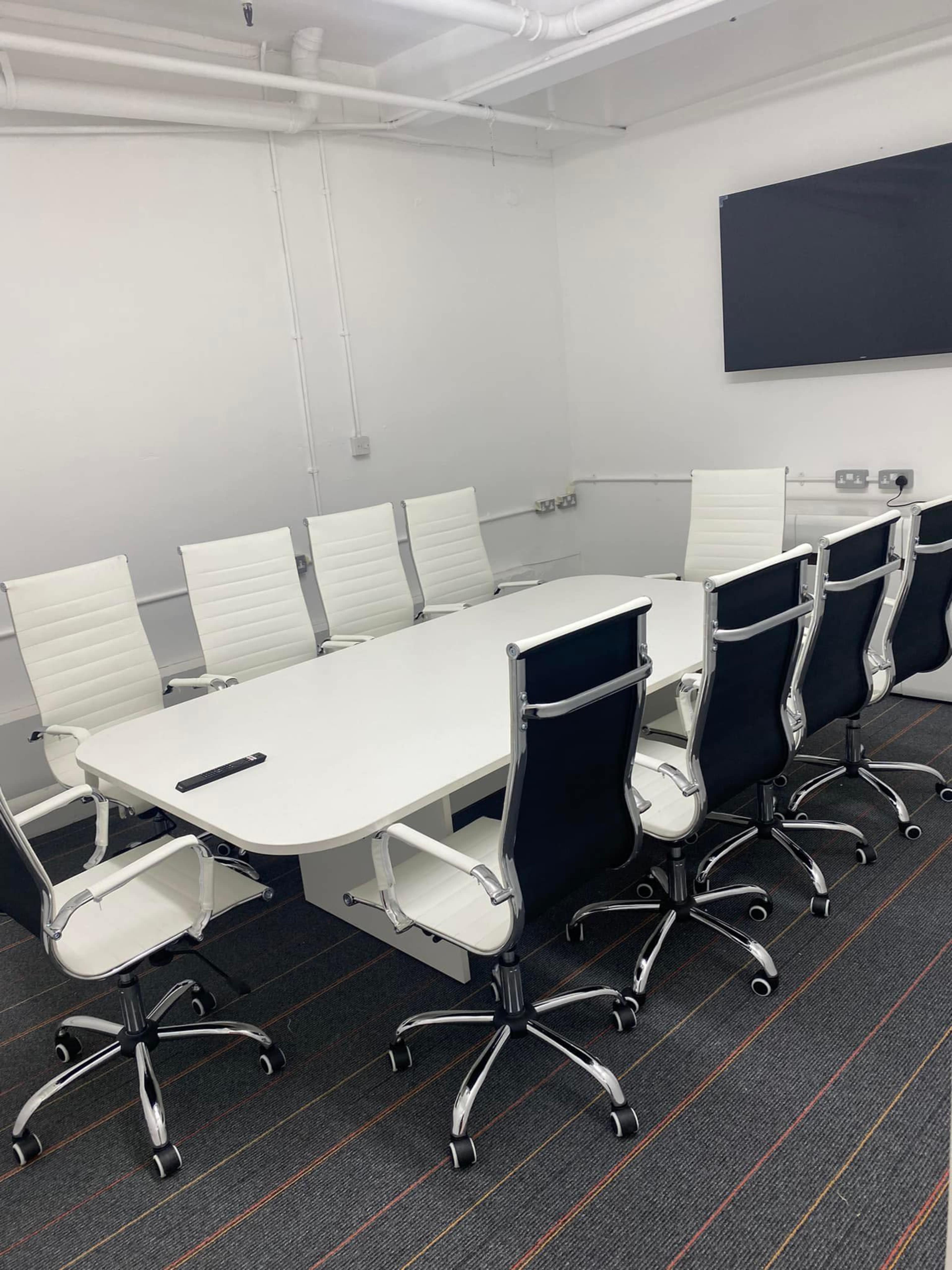 A modern conference room features a long white table surrounded by eight white and black office chairs, with a wall-mounted screen in the background.