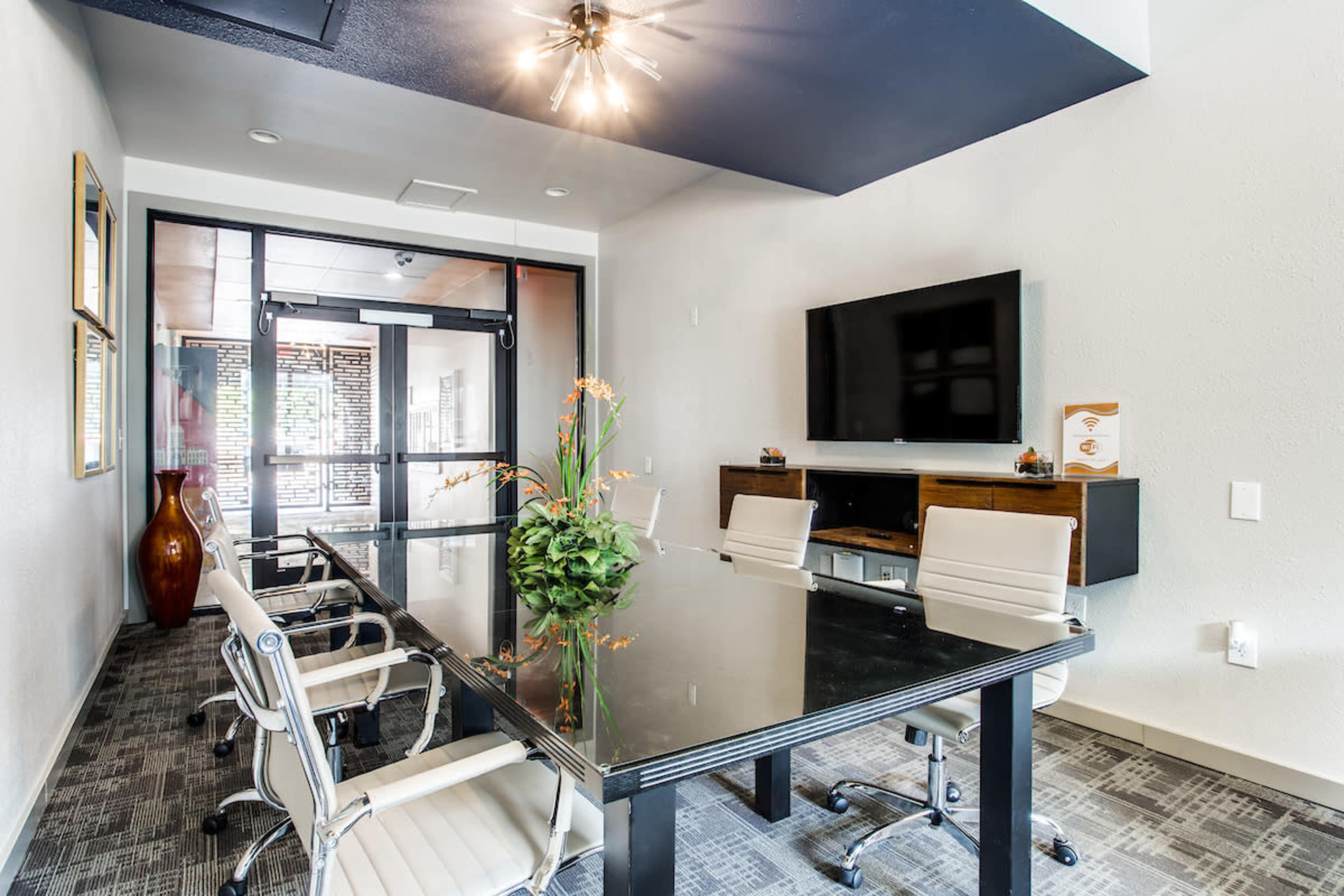 A modern conference room features a glass table surrounded by white chairs, with a television mounted on the wall and large windows allowing natural light.