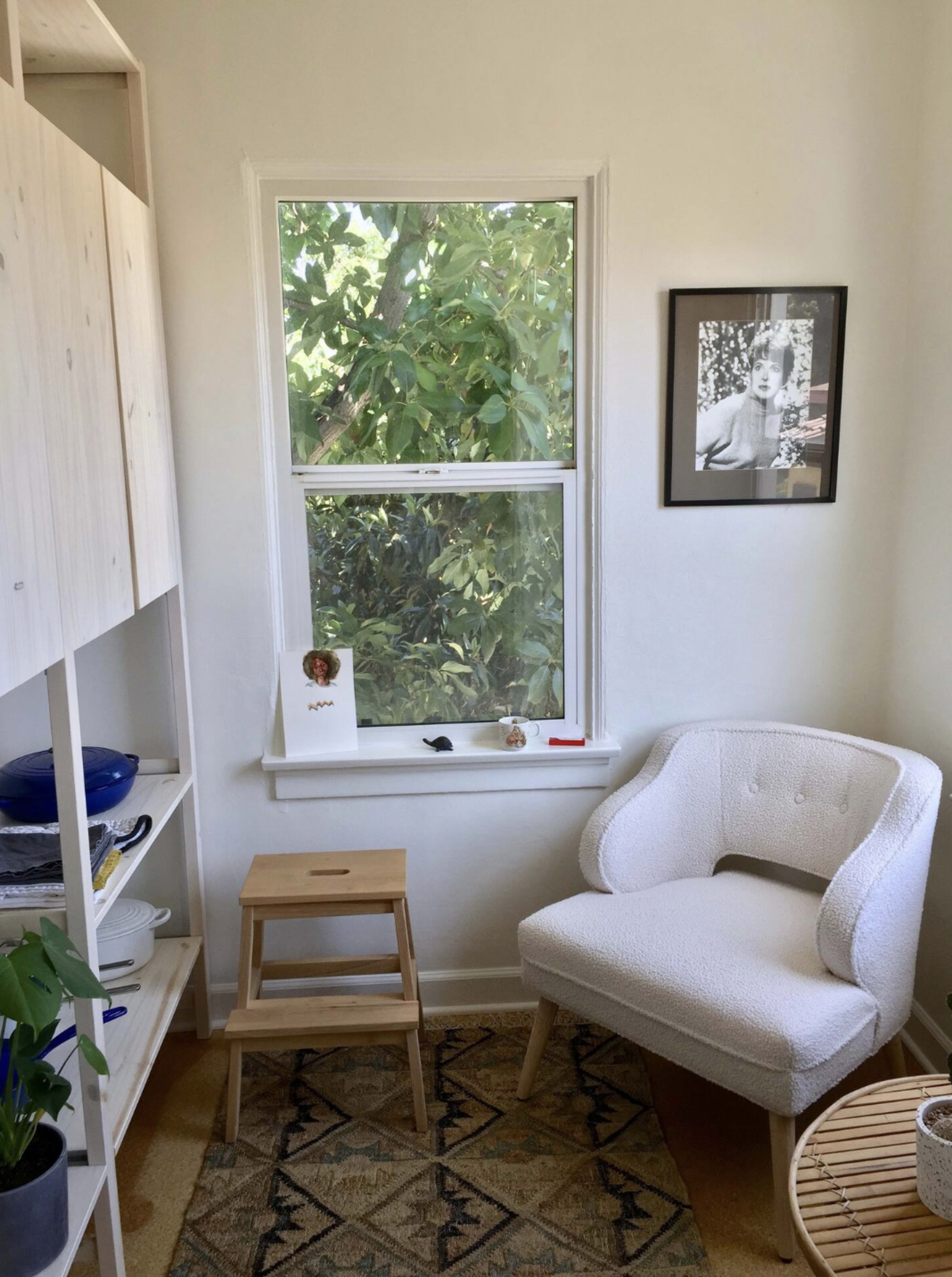 A cozy corner features a white chair, a small wooden stool, a bookshelf with blue dishes, and a window framed by greenery outside.