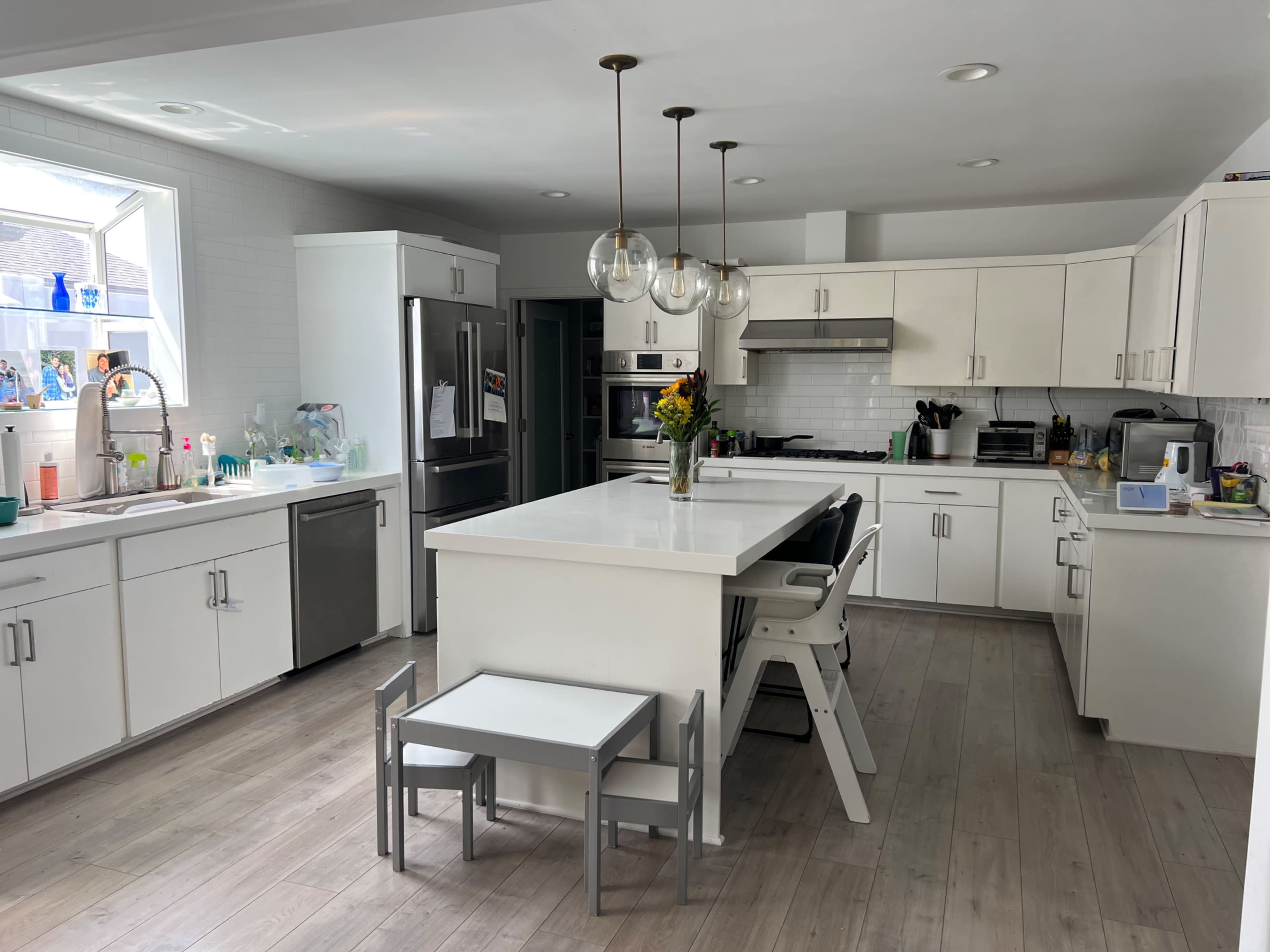 The image shows a modern kitchen with white cabinetry, a central island with seating, and large windows providing natural light.