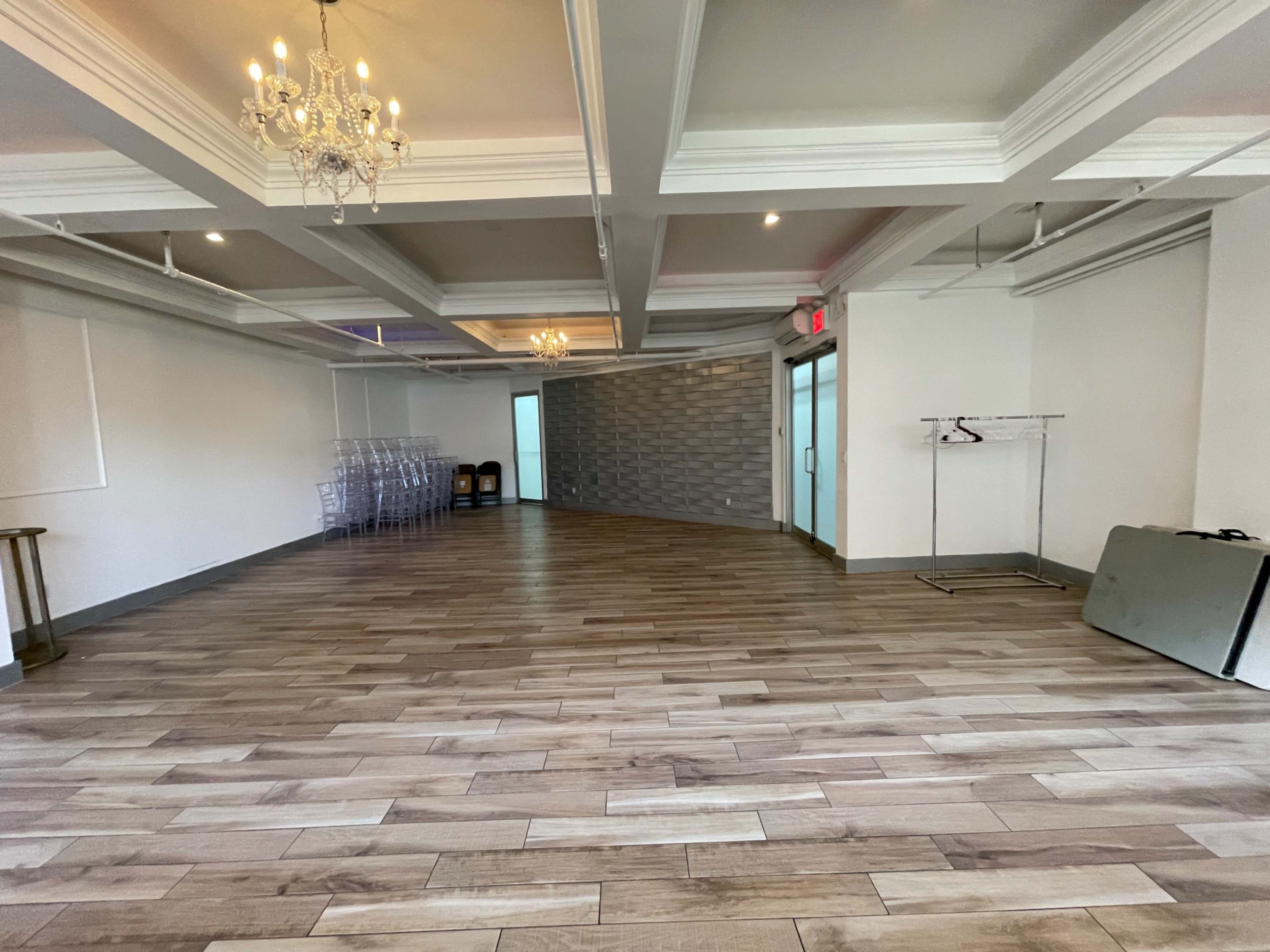 A spacious, empty room with wooden flooring, chandeliers, and a partitioned wall topped with decorative panels.