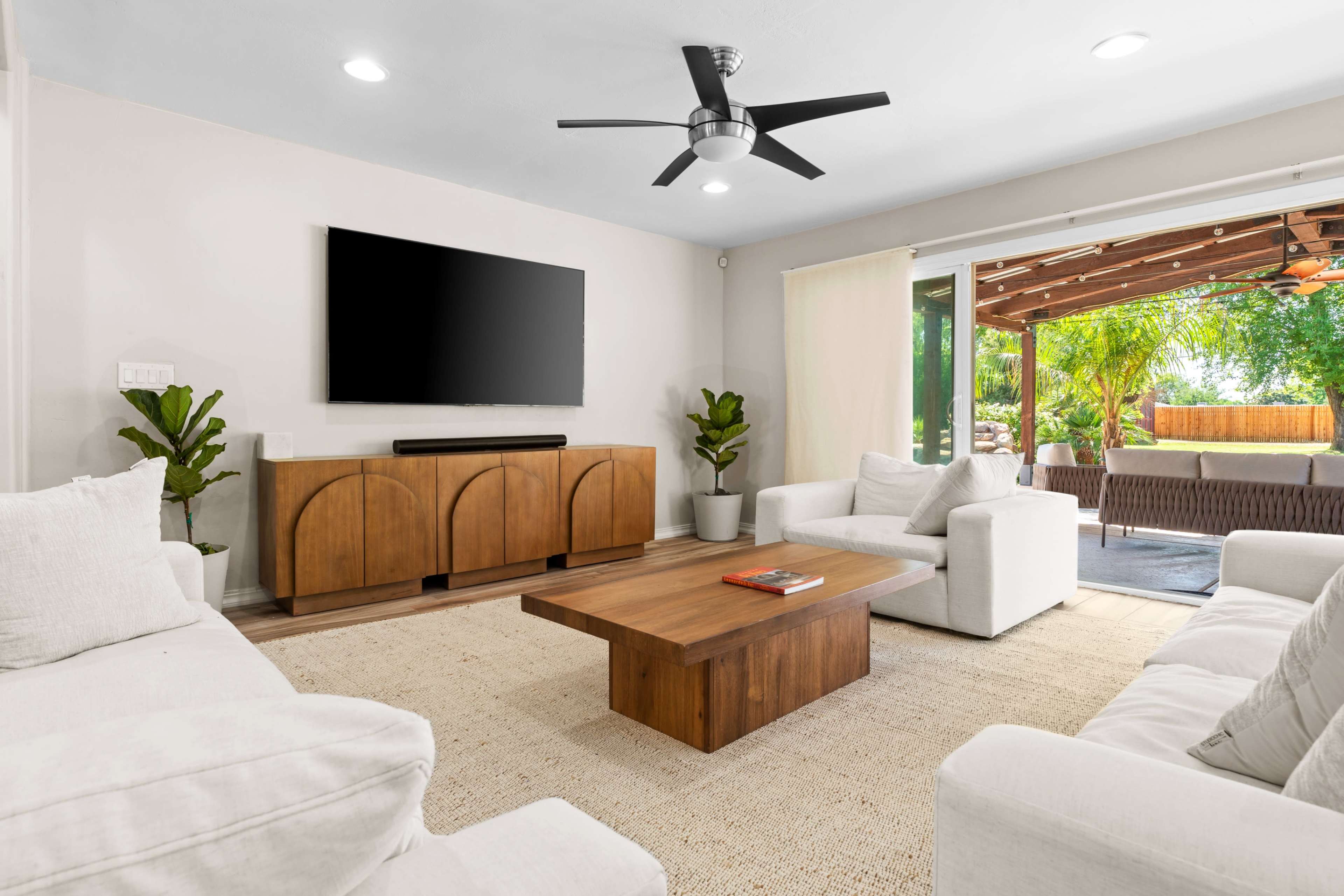 The living room features a modern layout with a large television mounted on the wall, a wooden coffee table, and white sofas arranged around a light area rug, with a view of an outdoor patio through sliding glass doors.