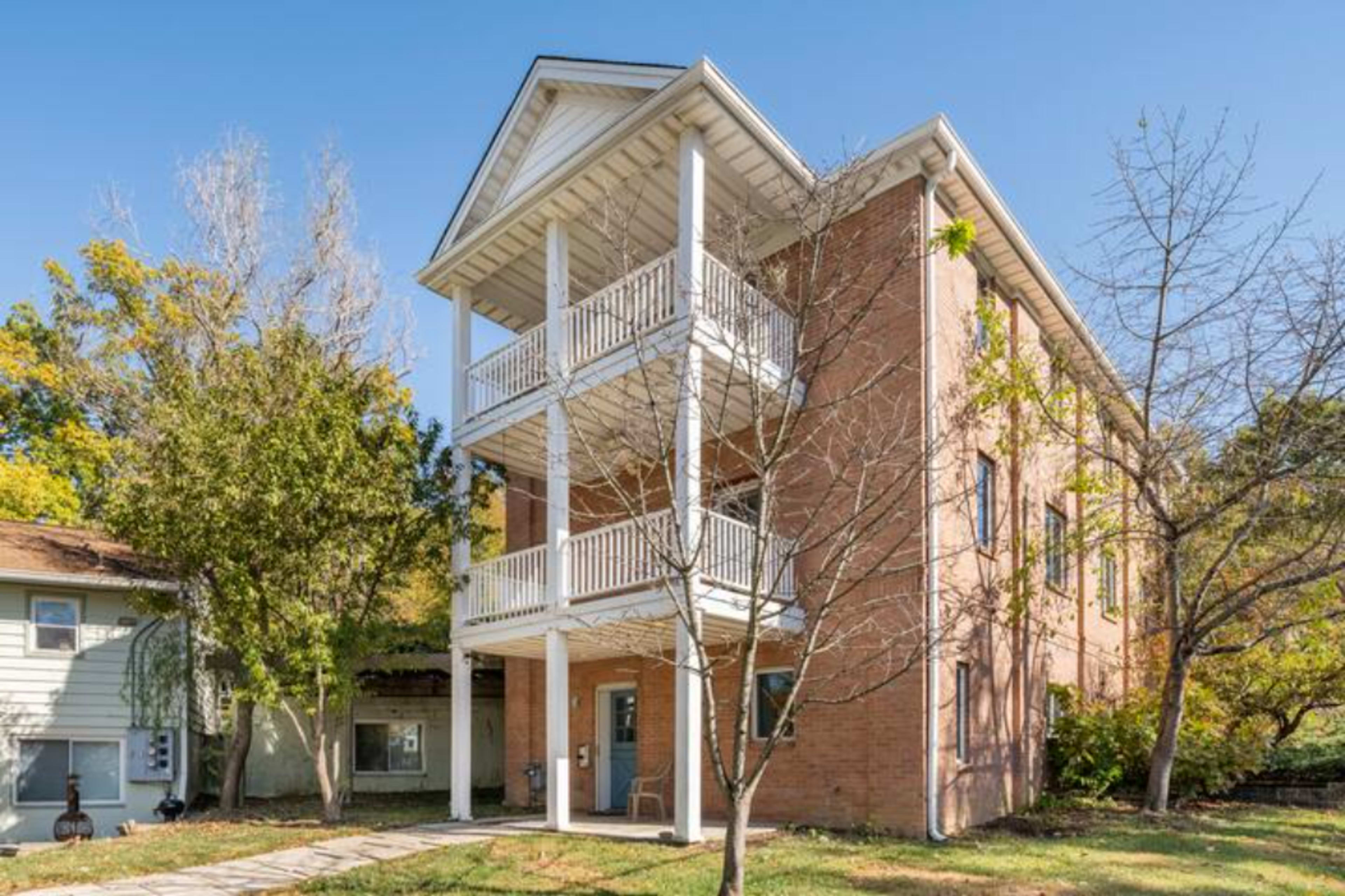 A three-story brick building with white balconies and a landscaped yard featuring trees.