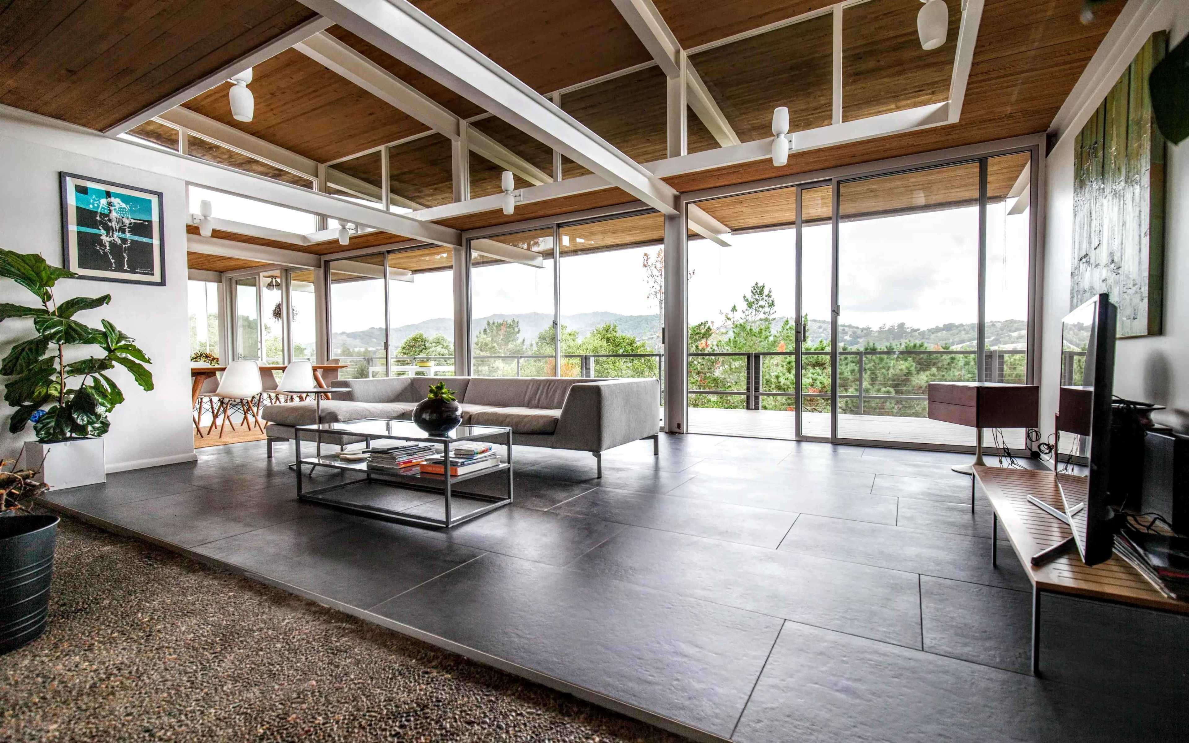 A modern living room with large windows, a gray sofa, a glass coffee table, and a view of a mountainous landscape.